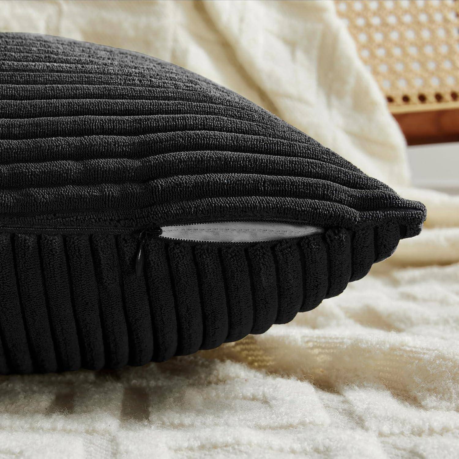 Ein schwarzes strukturiertes Kissen mit sichtbarem seitlichem Reißverschluss liegt auf einer beigen Decke auf einem Stuhl.