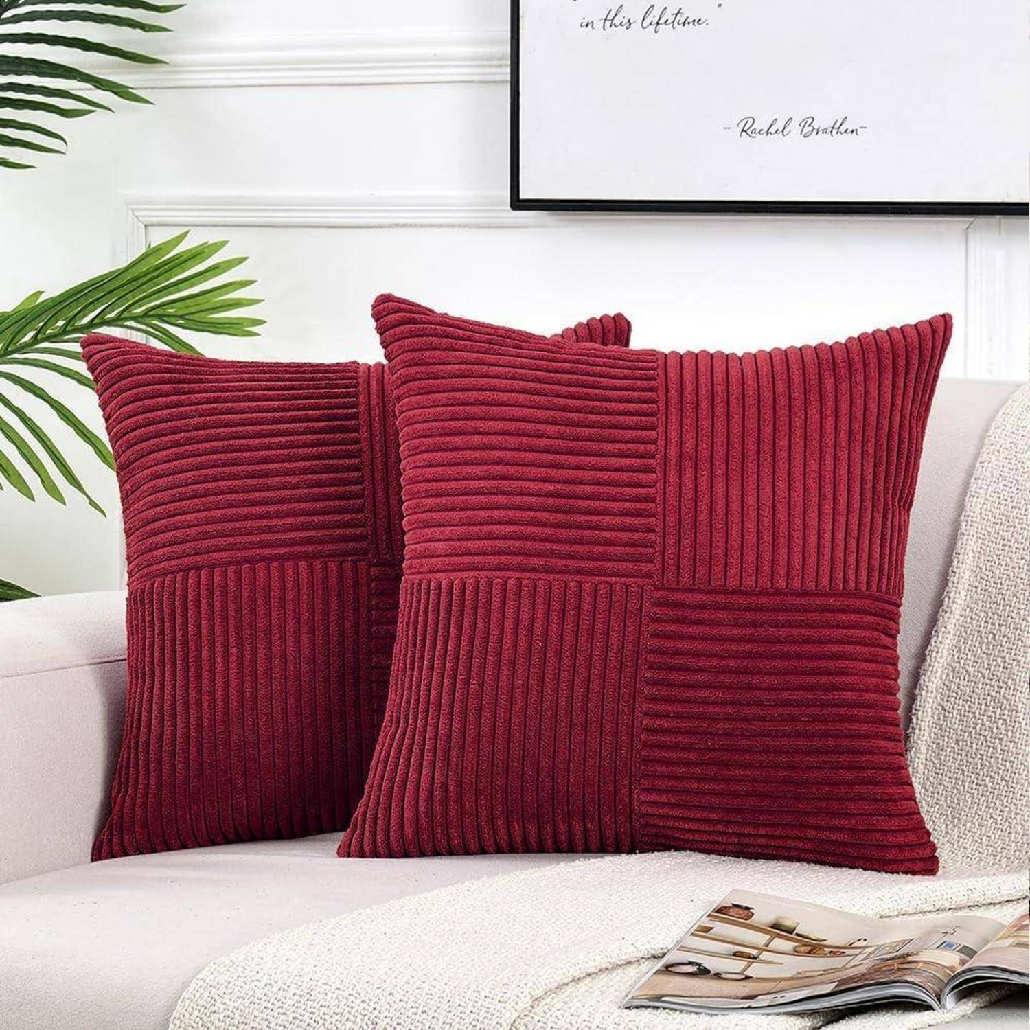 Zwei rote Kissen mit einem strukturierten Muster auf einem beigen Sofa, in der Nähe einer weißen Wand mit einer blattreichen Pflanze und einem gerahmten Bild darüber.