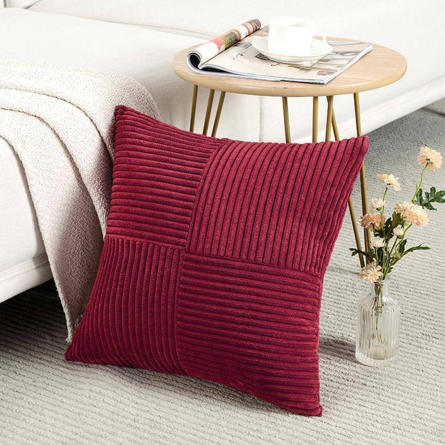 Ein gemütlicher Wohnbereich zeigt ein rotes strukturiertes Kissen auf einem cremefarbenen Sofa. In der Nähe steht ein kleiner Tisch mit einer Tasse und Zeitschriften, daneben befinden sich Blumen.
