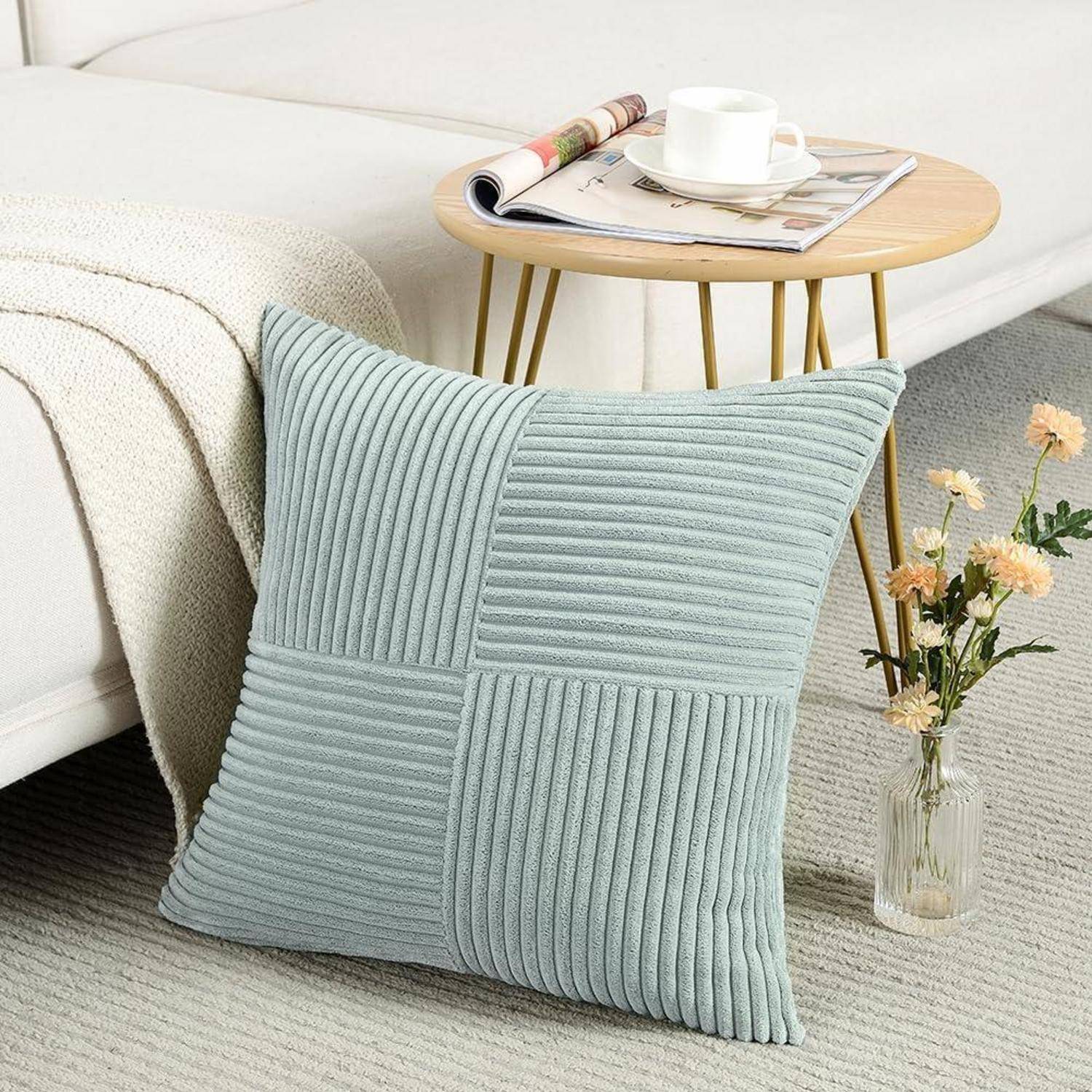Ein hellblau strukturiertes Kissen auf einem beigen Sofa, neben einem kleinen runden Holztisch mit einer Zeitschrift und Blumen in einer Vase.