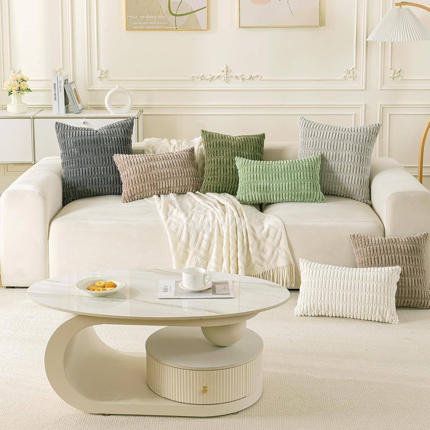Ein gemütliches Wohnzimmer mit einem weißen Sofa, das mit unterschiedlichen Zierkissen und einer weichen Decke geschmückt ist. Ein moderner Couchtisch trägt einen kleinen Teller.