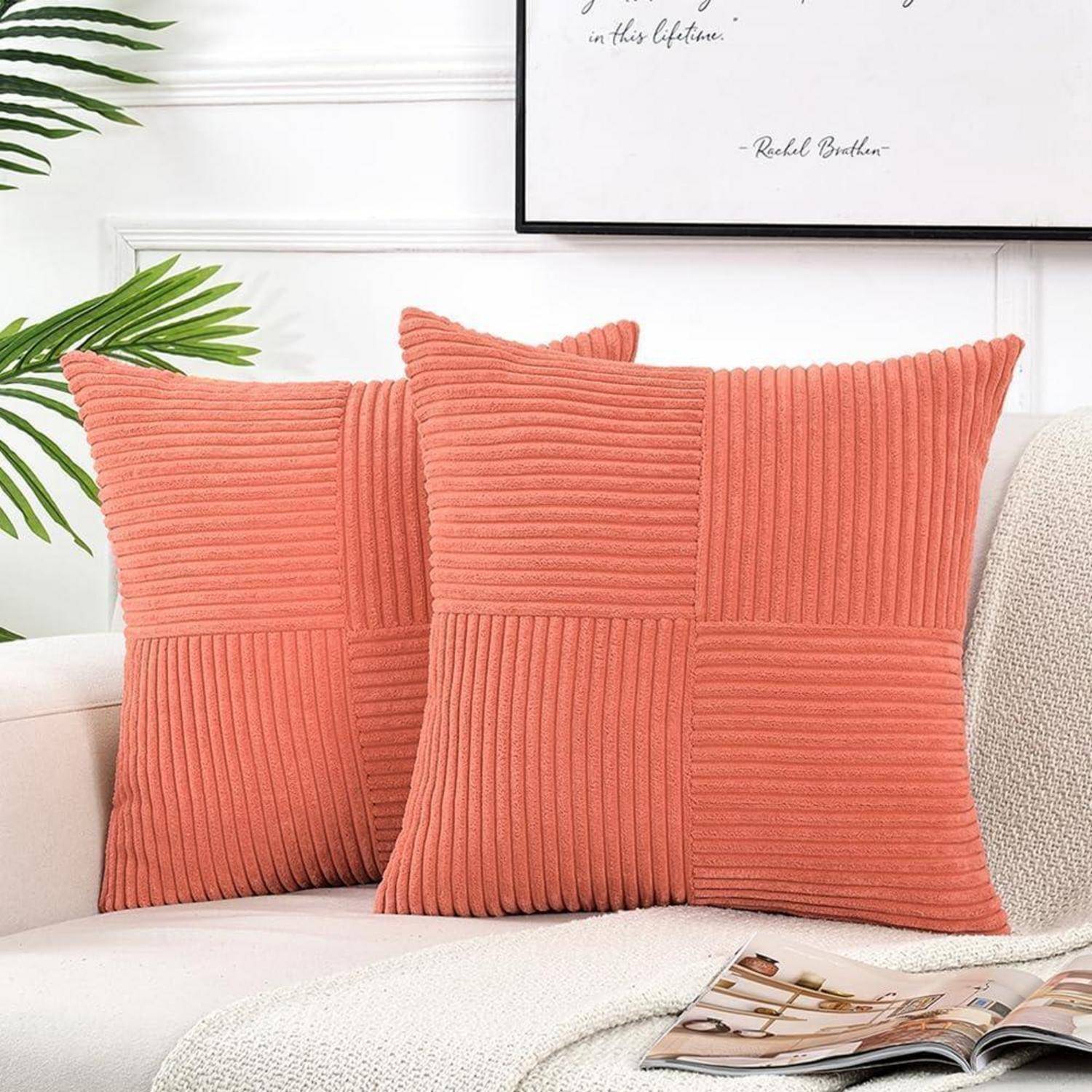 Zwei orangefarbene strukturierte Kissen auf einem weißen Sofa, mit einem gerahmten Zitat an der Wand darüber, umgeben von grünen Blättern, eine gemütliche Atmosphäre schaffend.