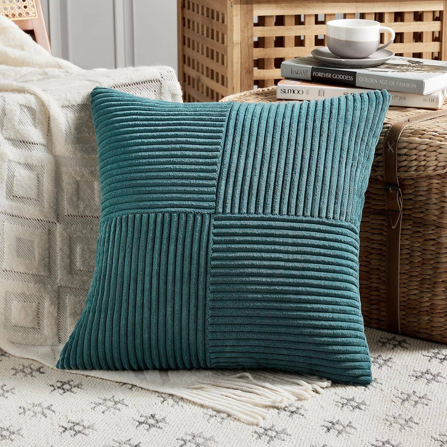 Ein türkisfarbenes gestreiftes Kissen ruht auf einem beigen gemusterten Sofa; daneben befinden sich ein Flechtkorb mit einer Teetasse, drei Büchern und einer grauen Decke.