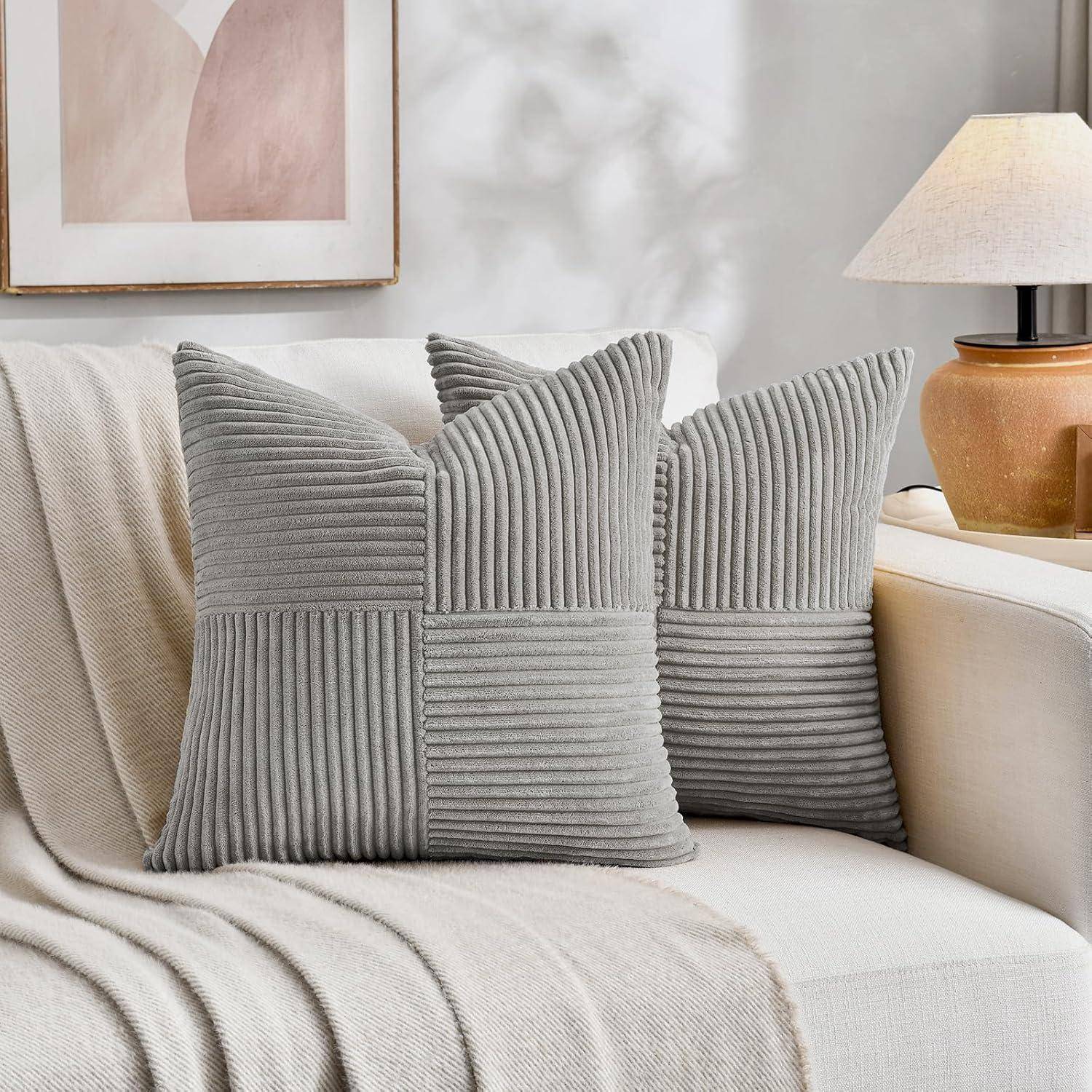 Zwei gerippte, graue Kissen auf einem hellen Sofa mit einer beigen Decke, neben einer Lampe und abstrakter Kunst an einer hellgefärbten Wand.
