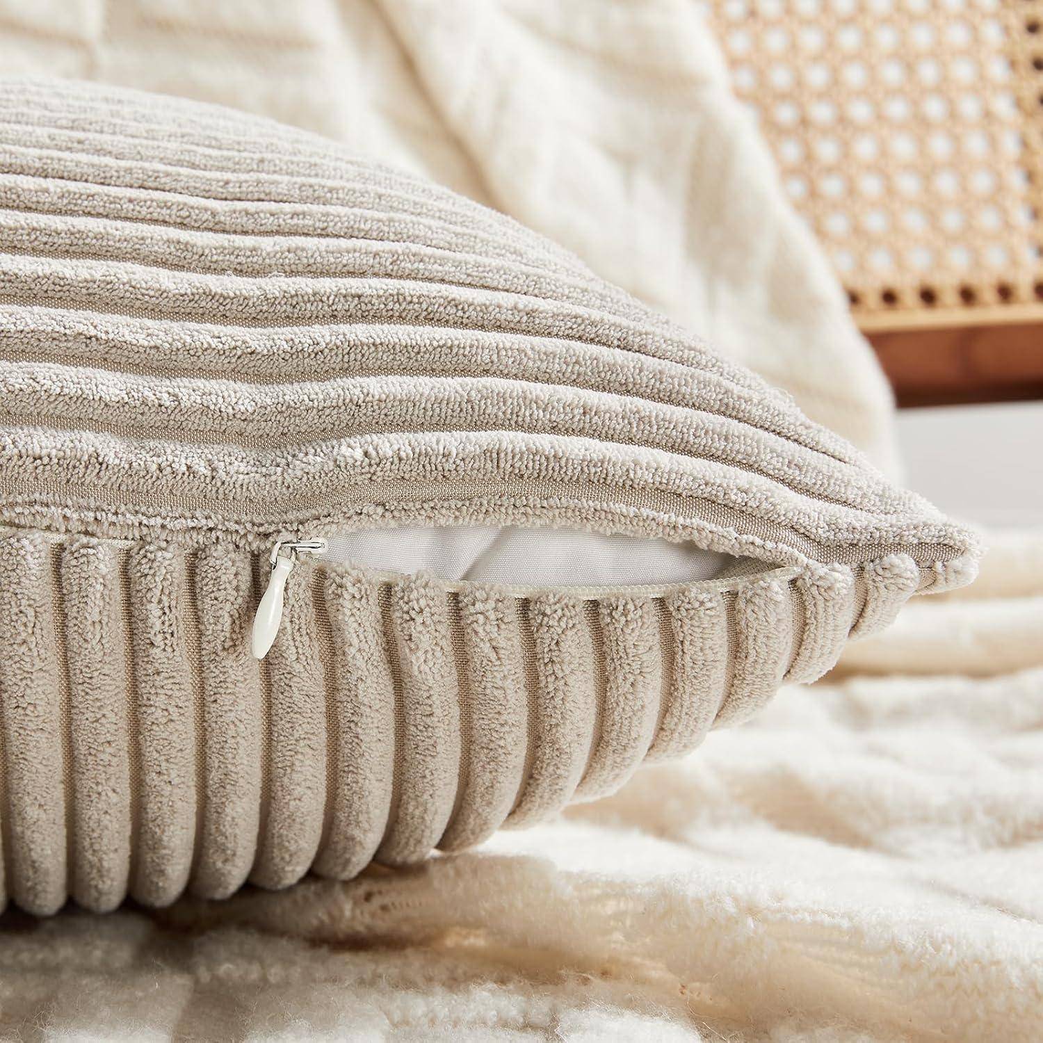 Ein plüschiges, geripptes beiges Kissen mit sichtbarem Reißverschluss ruht auf einer weichen, beigen strukturierten Decke. Ein Holzstuhl mit Geflecht-Rückenlehne ist teilweise zu sehen.