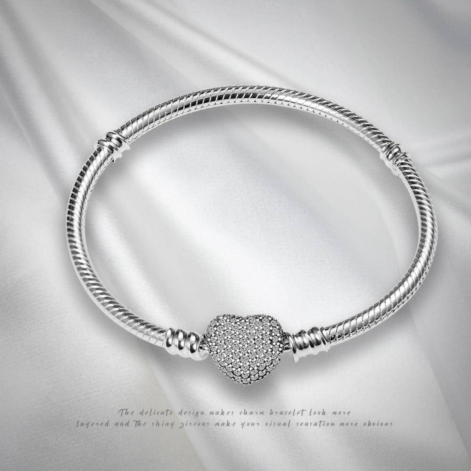 Silberarmband mit einem strukturierten herzförmigen Mittelelement, das auf seidigem Stoff platziert ist. Eine dezente Beschriftung darunter liest: „Das feine Design macht das Charm-Armband geschichteter aussehen..."