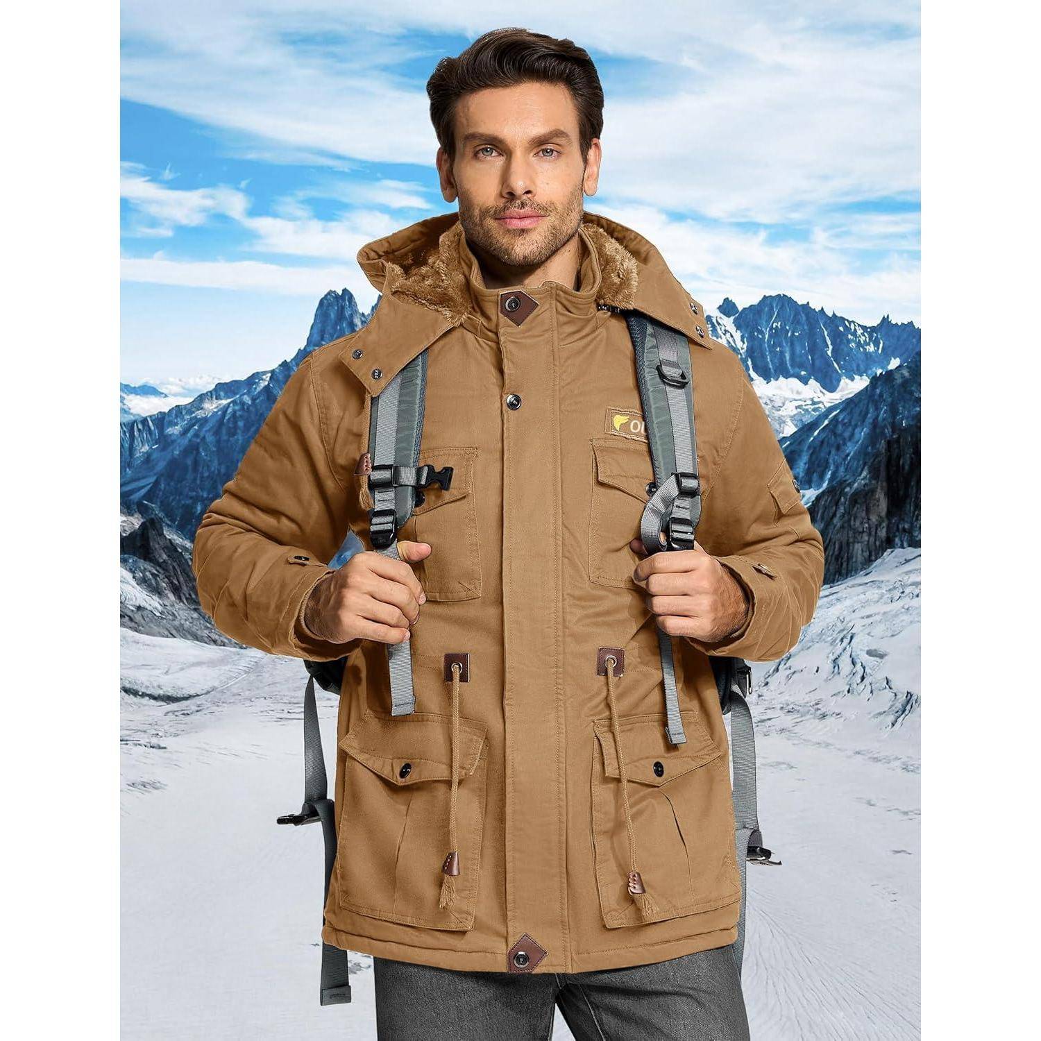 Eine Person steht auf einem schneebedeckten Bergpfad, trägt einen beigen Wintermantel und einen Rucksack, mit rauen Berggipfeln im Hintergrund unter einem blauen Himmel.