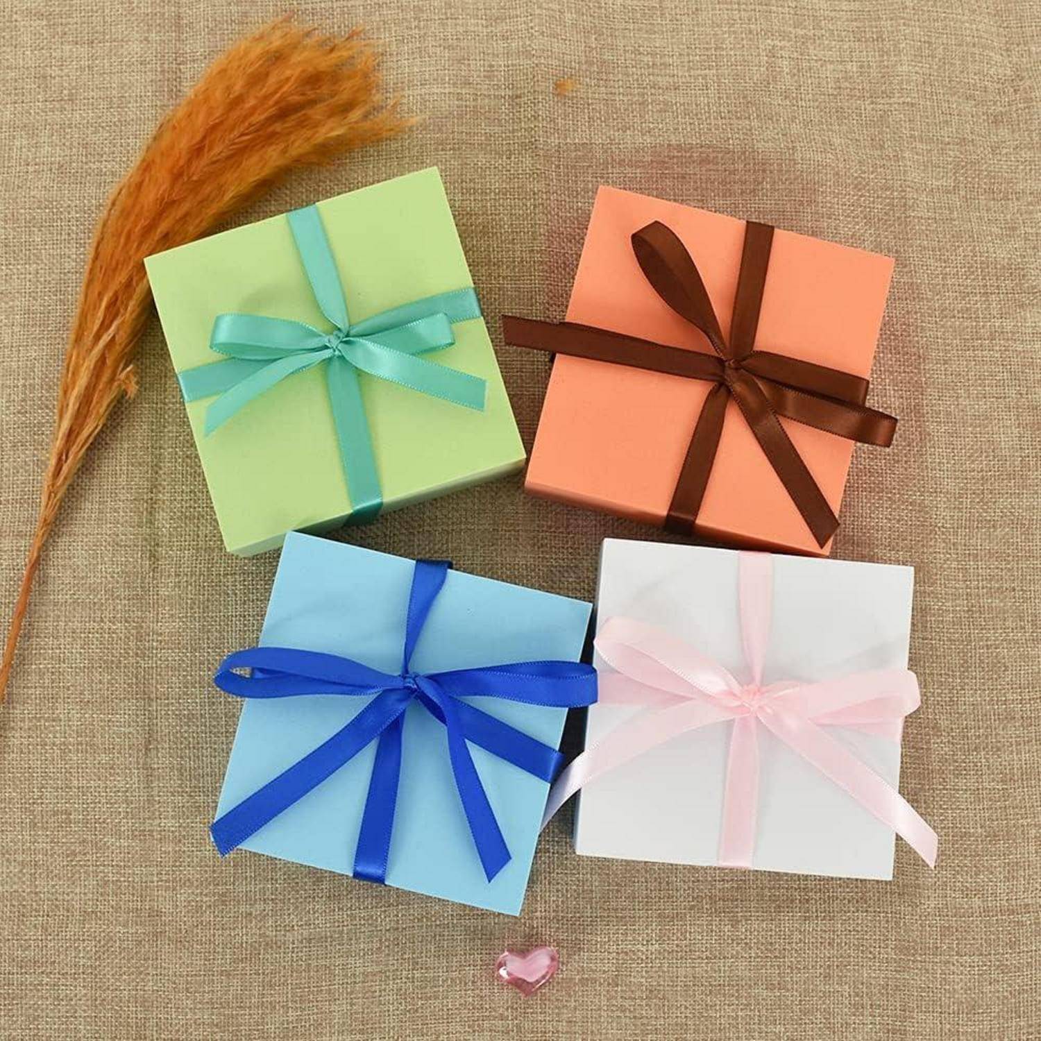 Vier Geschenkkartons mit farbigen Bändern (grün, orange, blau und rosa) sind auf einer Juteoberfläche platziert. Daneben liegt ein getrockneter Weizenhalm.