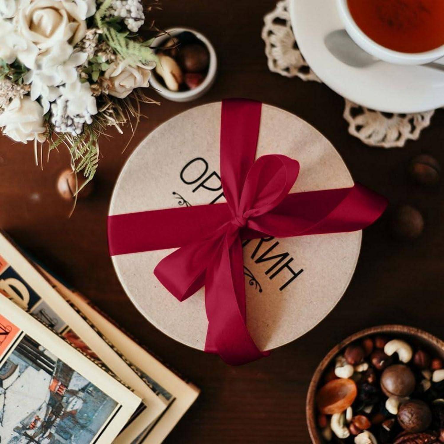 Eine runde Geschenkbox mit einem roten Band liegt auf einem Tisch neben einer Tasse Tee, einer Schale mit gemischten Nüssen und einem Blumenarrangement.