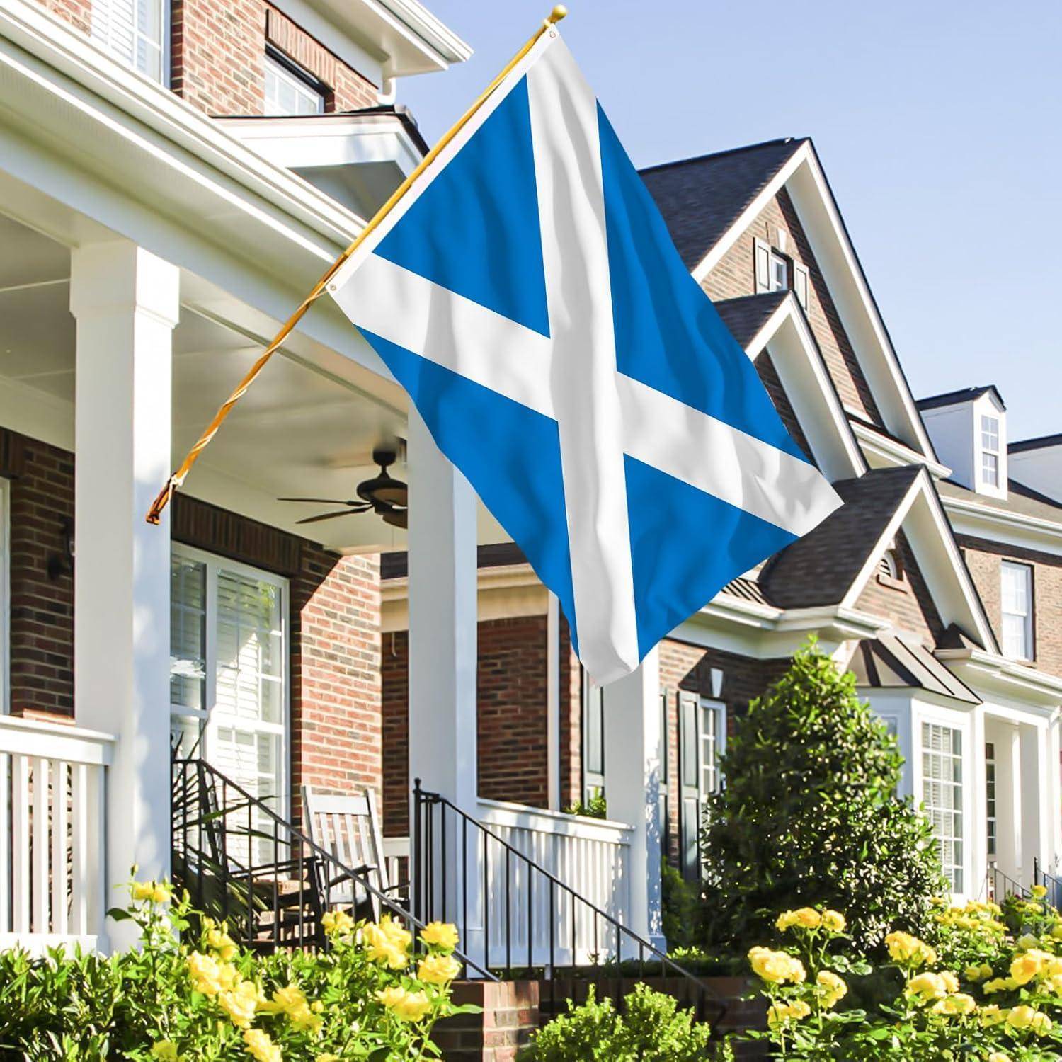 Eine Andreaskreuz-Flagge, die Schottland symbolisiert, weht an einem Mast vor einer Reihe von roten Ziegelhäusern mit weißer Einfassung und Grünbepflanzung.