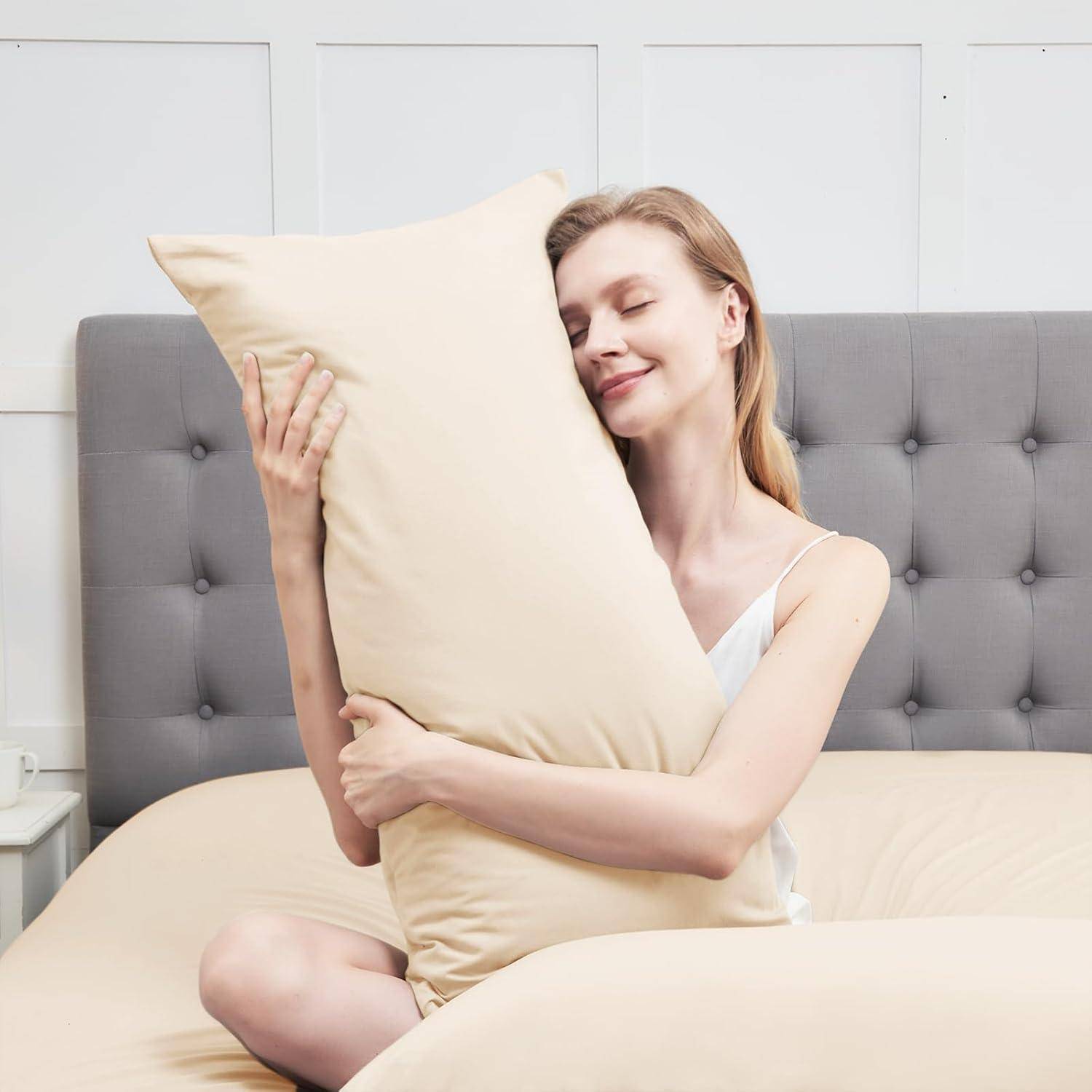 Eine Person sitzt auf einem Bett, lächelt und umarmt ein großes Kissen. Die Umgebung ist ein Schlafzimmer mit einem grauen Kopfteil und weißer Bettwäsche.