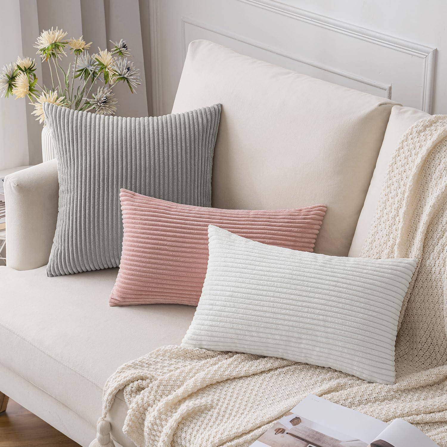 Drei strukturierte Kissen in Grau, Rosa und Weiß sind auf einem weißen Sofa mit einer beigen Strickdecke angeordnet. Eine Vase mit Blumen befindet sich im Hintergrund.
