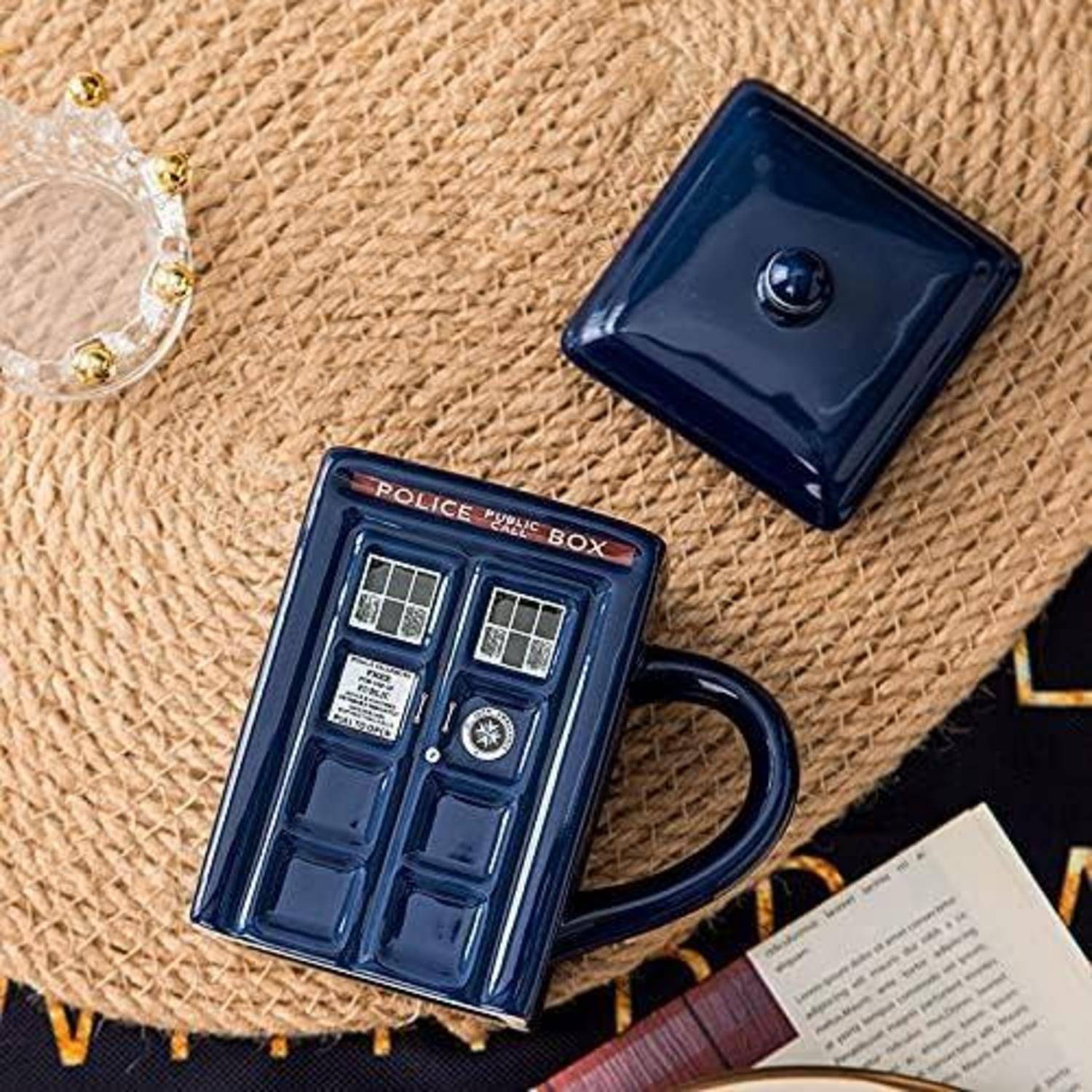Eine TARDIS-thematische blaue Tasse mit Deckel steht auf einer gewebten Matte neben einem aufgeschlagenen Buch und einem dekorativen Glasgegenstand.