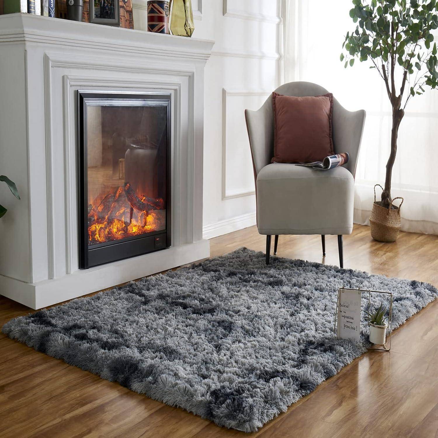 Ein gemütlicher Sitzbereich zeigt einen beleuchteten Kamin neben einem grauen Sessel mit einem Kissen. Ein plüschiger grauer Teppich liegt auf Holzfußboden.