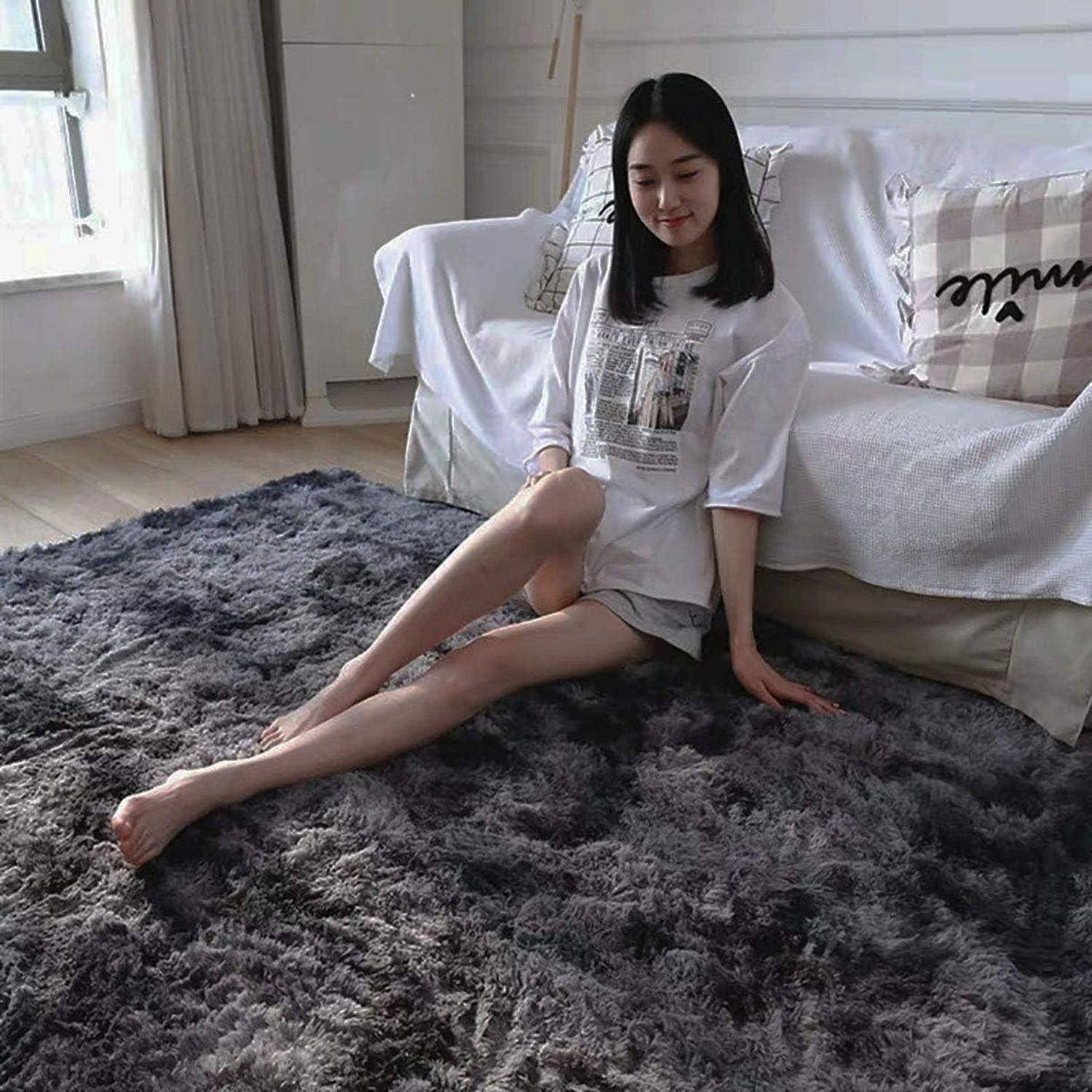 Eine Person, die auf einem grauen Teppich in einem Wohnzimmer sitzt und einen lässigen Outfit trägt. Im Hintergrund befindet sich ein Sofa mit einem gemusterten Kissen.