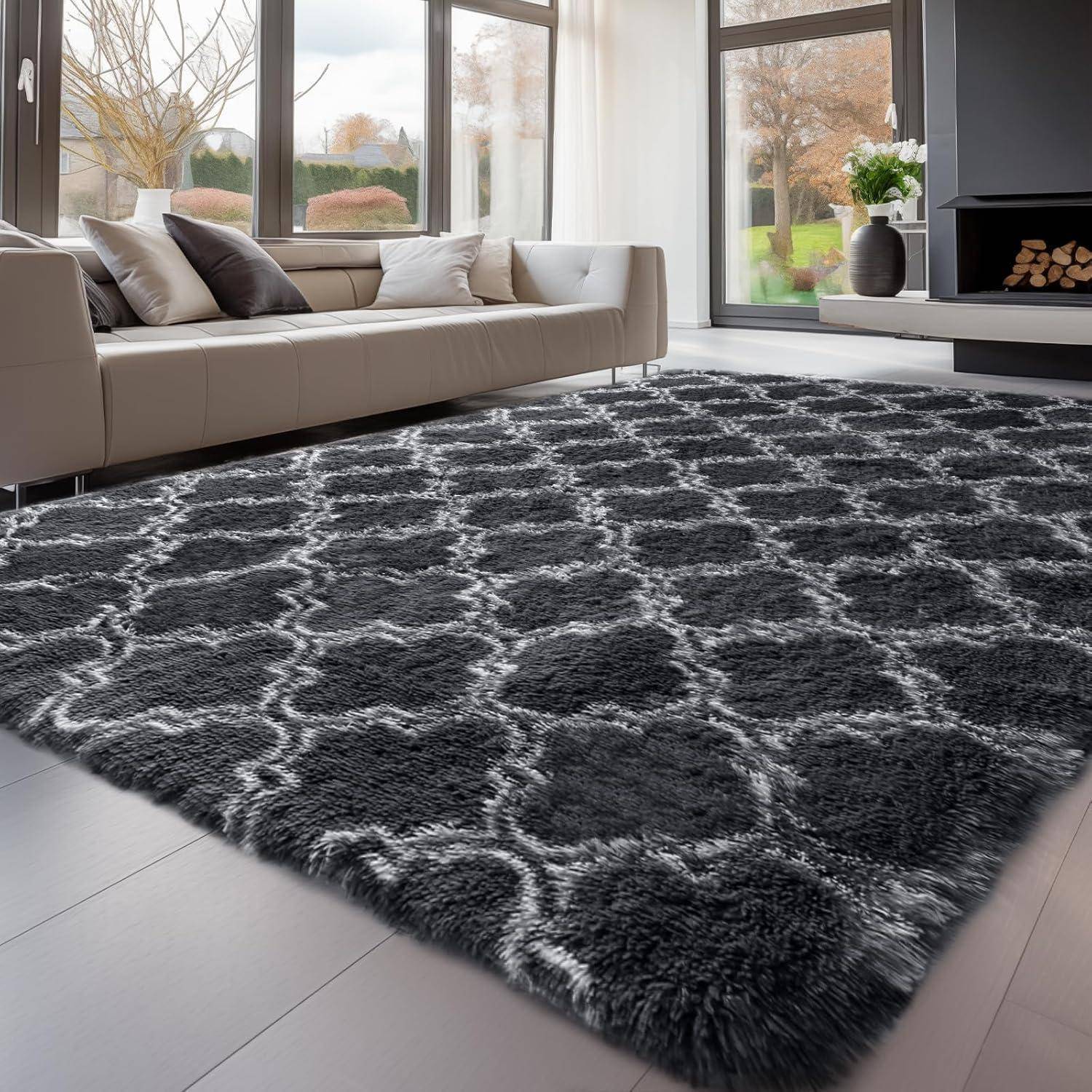Ein gemütliches Wohnzimmer mit einem großen, gemusterten grauen Teppich auf einem Holzboden. Ein Ecksofa mit grauen und weißen Kissen steht neben großen Fenstern.