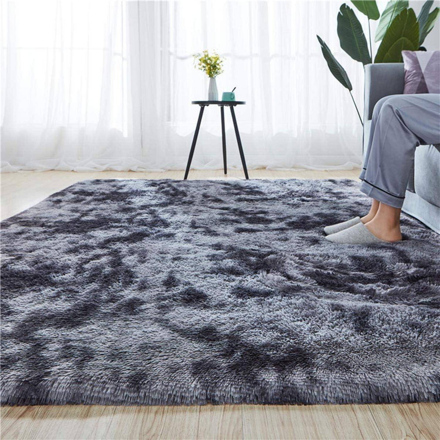 Ein plüschiger grauer Teppich in einem Wohnzimmerambiente mit einem kleinen runden Tisch, auf dem eine Vase steht. Eine Person auf einem Sofa ist teilweise sichtbar.
