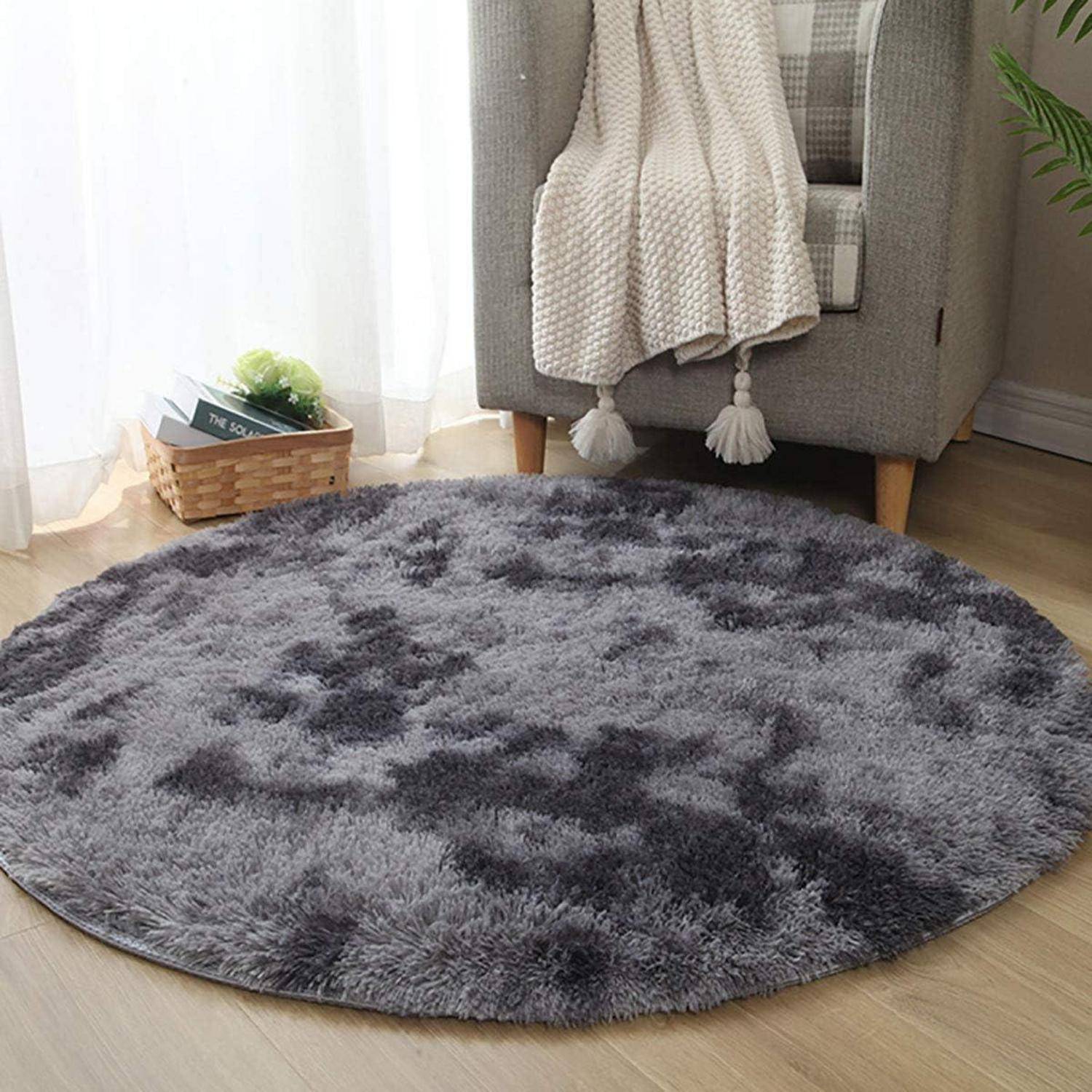 Ein rundes, graues Shaggy-Teppich, der in einem gemütlichen Raum mit einem beigen Sessel und einer leichten Decke, die darüber drapiert ist, neben einem kleinen Korb platziert ist.