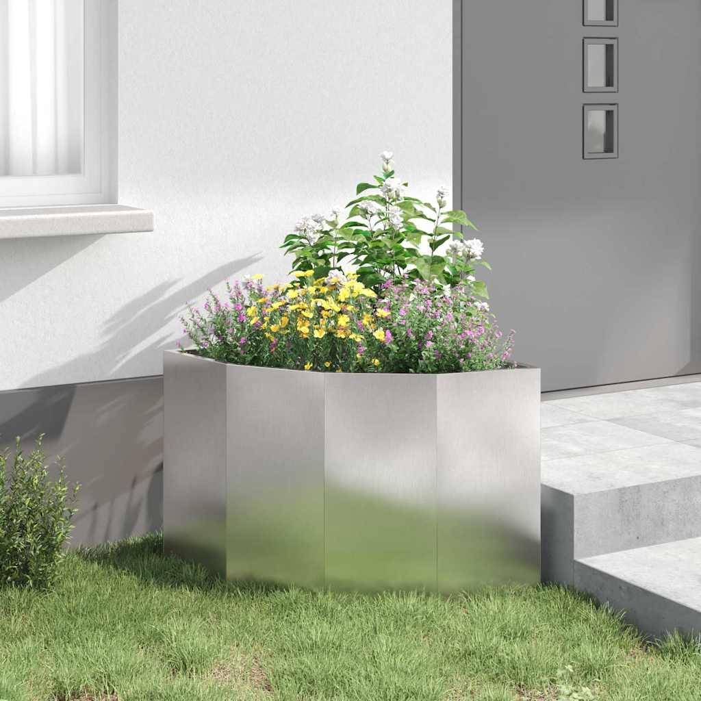 Ein Pflanzgefäß aus Metall mit leuchtenden Blumen und grünem Blattwerk steht auf einer Betonterrasse neben der Wand eines modernen Gebäudes und einem Grasstück.