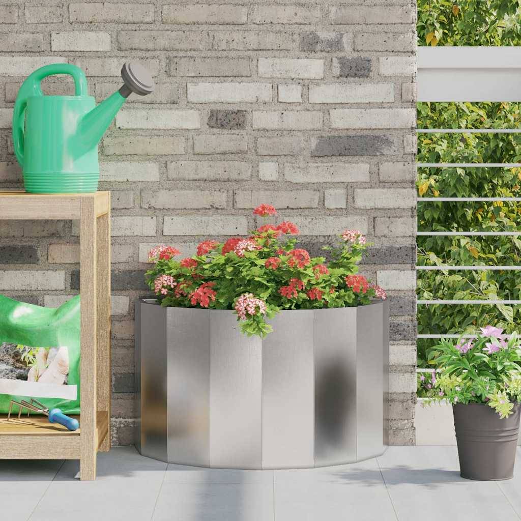 Ein Metallpflanzkübel mit roten und rosa Blumen auf einer Terrasse, neben einer Holzbank, auf der eine grüne Gießkanne steht.
