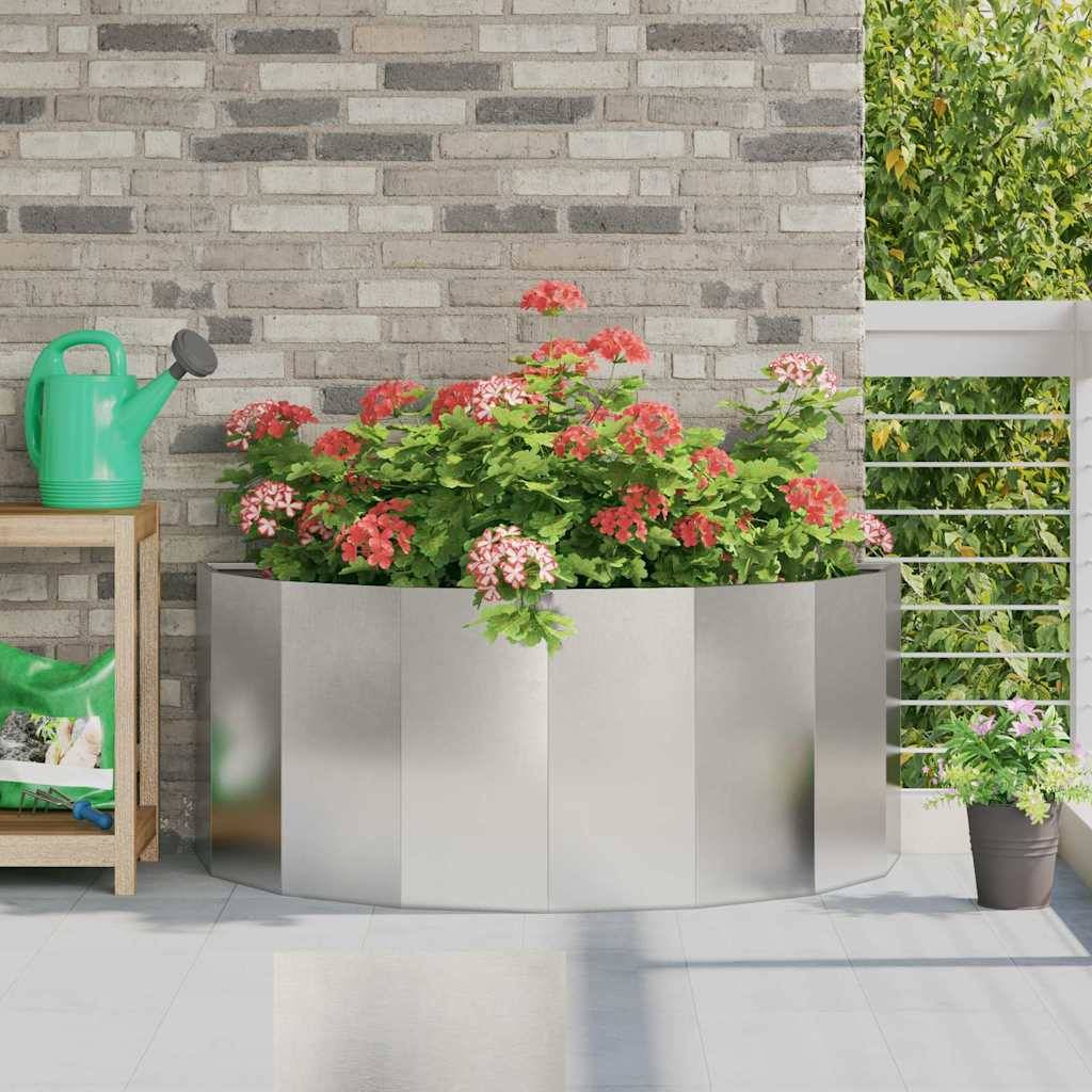 Ein reflektierender Metallpflanzer mit rosa Blumen steht auf einem Balkon mit Ziegelwand im Hintergrund. Eine Gießkanne und ein Topfpflanze befinden sich in der Nähe.