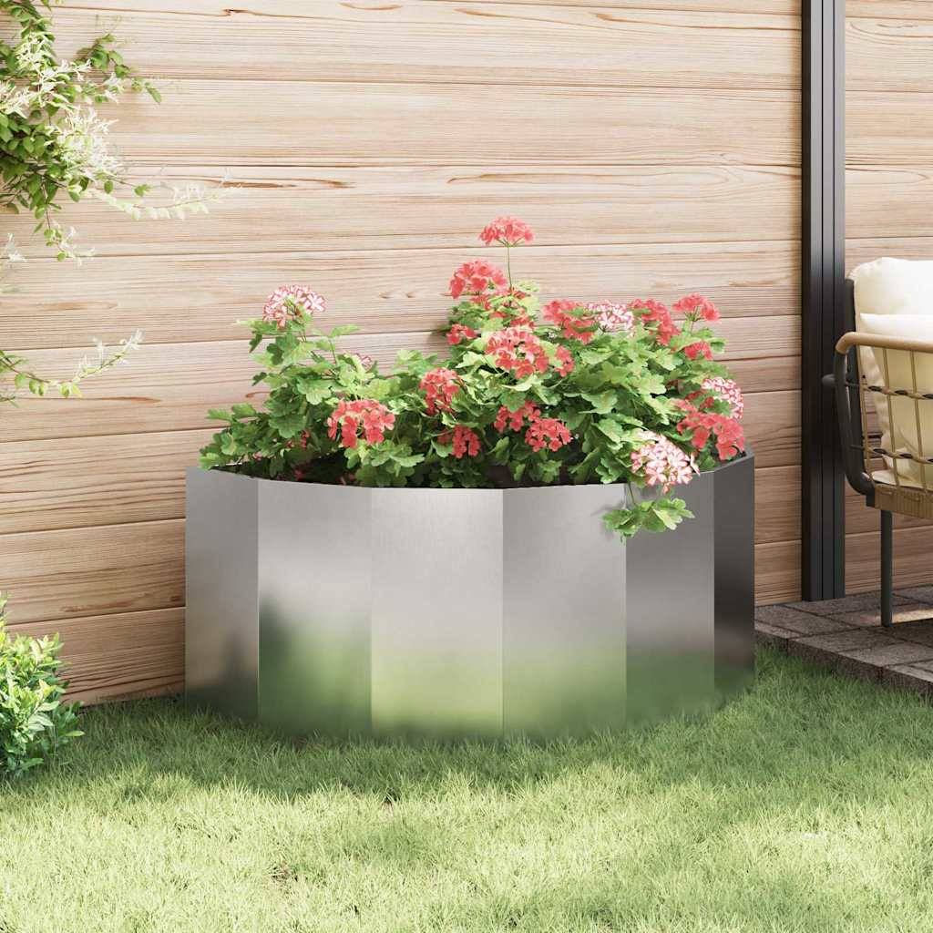 Ein sechseckiger Metallpflanzkübel, gefüllt mit blühenden roten Begonien, steht auf einer Terrasse vor einer Holzwand, mit Grünpflanzen im Hintergrund.