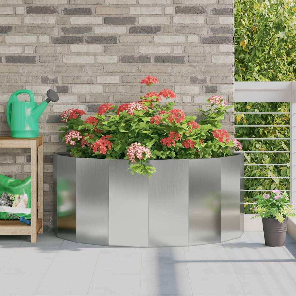 Ein Metallpflanzer mit blühenden roten und rosa Blumen steht auf einer Terrasse, umgeben von einer Gießkanne und einer kleinen Holzbank.