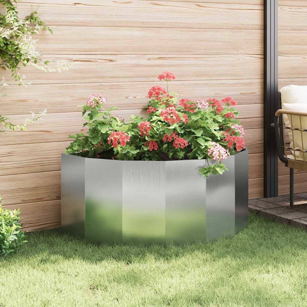 Ein Metallpflanzer mit blühenden roten Blumen steht auf grünem Gras vor einer Holzwand, links daneben ein kleiner Baum.