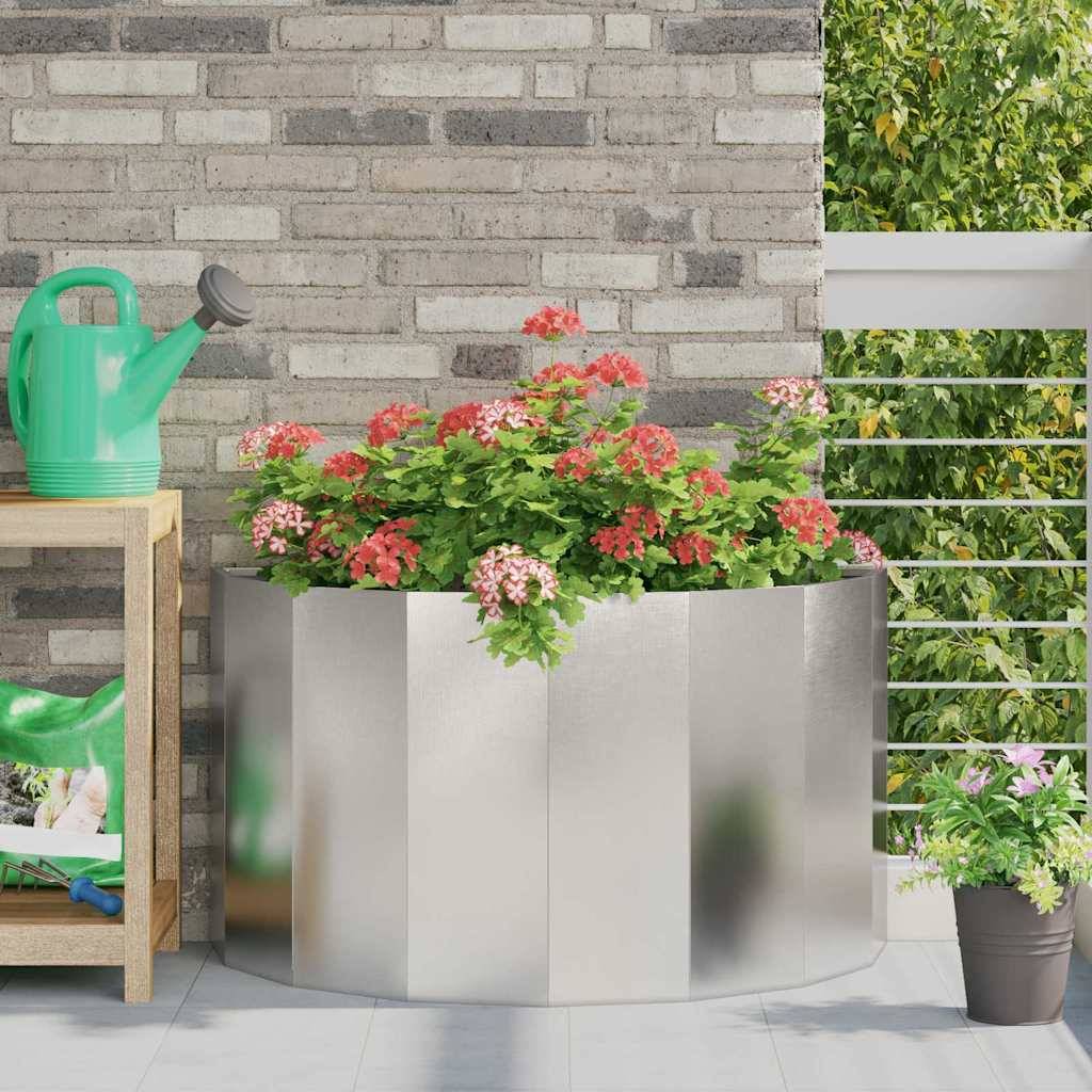 Ein Pflanzkübel aus Edelstahl mit blühenden rosa Blumen steht auf einer Terrasse. In der Nähe befinden sich eine grüne Gießkanne und ein Regal mit Gartengeräten.