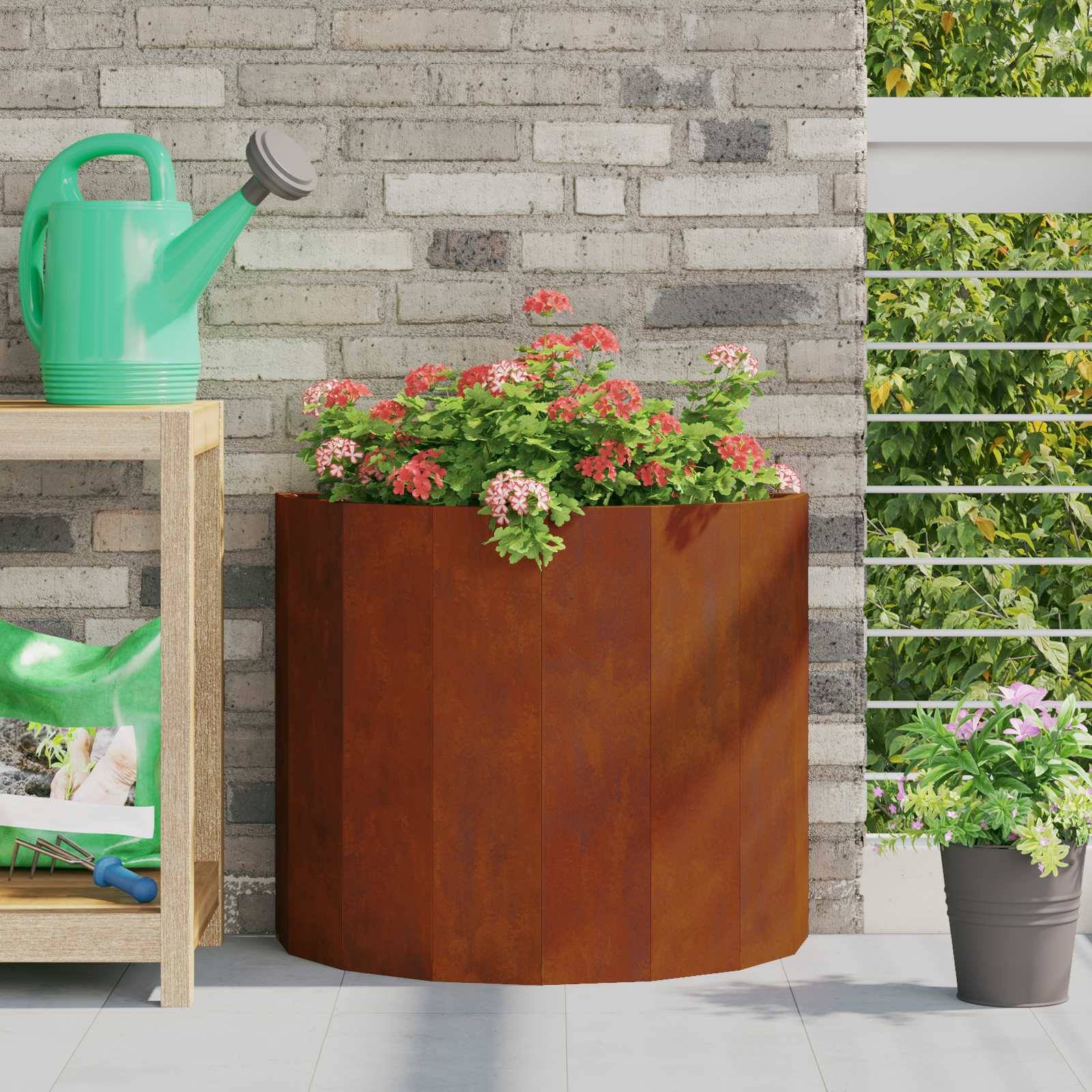 Ein Holzregal mit einer grünen Gießkanne und Gartenbedarfsartikeln steht neben einem großen Blumenkübel mit blühenden rosa Blumen auf einer sonnigen Terrasse.