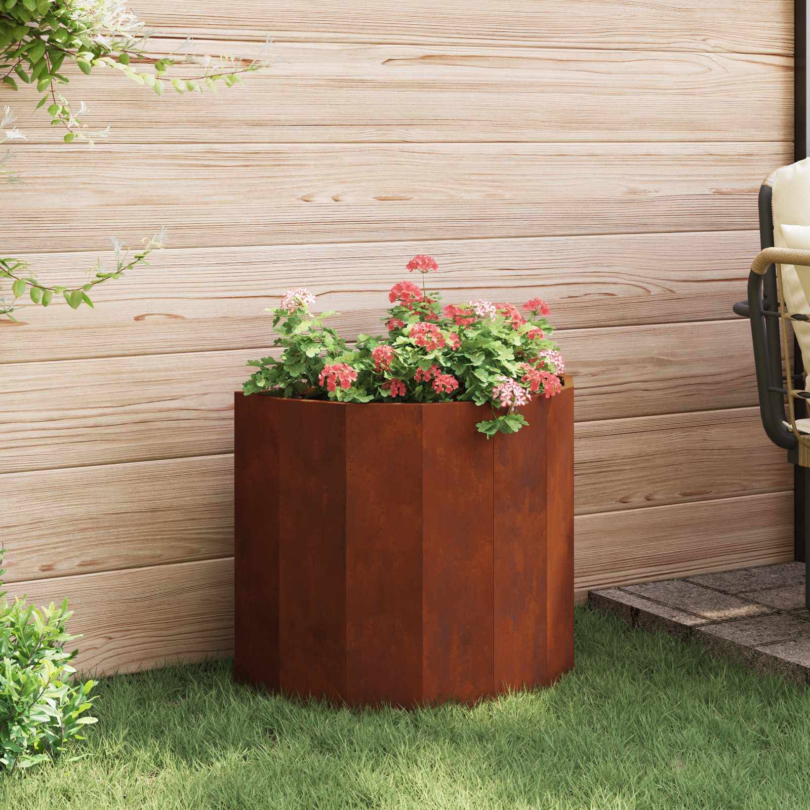 Ein hölzerner Blumenkübel gefüllt mit rosa Blüten und grünem Laub steht auf einem üppigen Rasen vor einer hellen Holzwand.