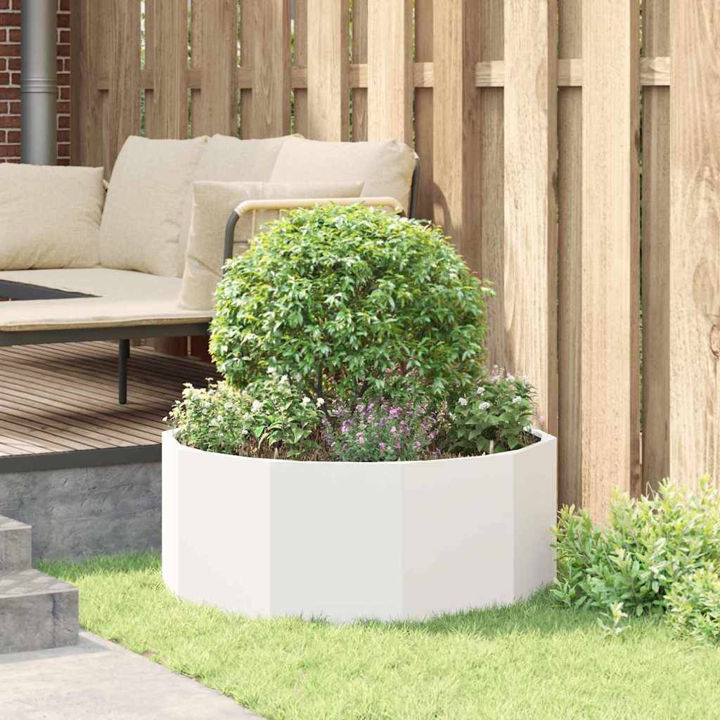 Ein runder, weißer Pflanzkübel mit üppigen grünen Sträuchern steht auf Gras neben einem Holzzaun. Eine Terrasse mit hellen Sitzmöbeln befindet sich im Hintergrund.