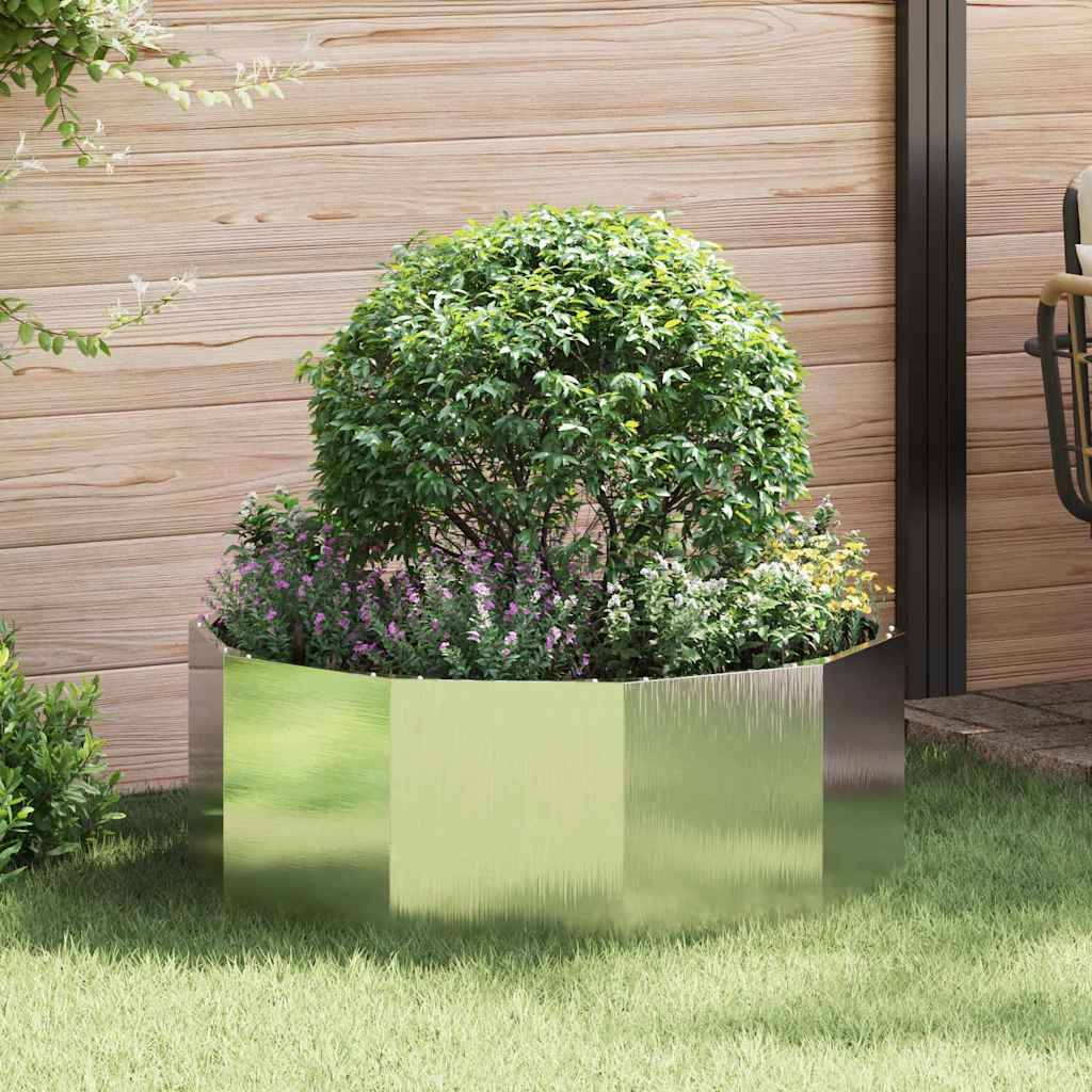 Ein metallischer, sechseckiger Pflanzkasten beherbergt einen kleinen, runden Strauch, umgeben von lila Blumen. Er steht vor einem Holzzaun in einer Gartenlandschaft.