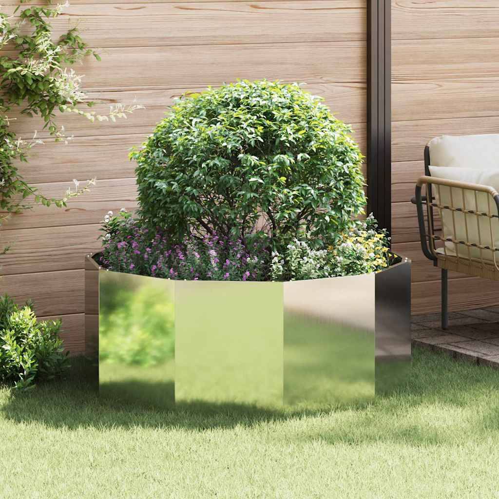 Ein üppiger grüner Busch mit kleinen Blüten in einem glänzenden, metallischen Pflanzkübel auf einer Terrasse. Daneben steht ein Stuhl, was auf eine Gartenumgebung hindeutet.