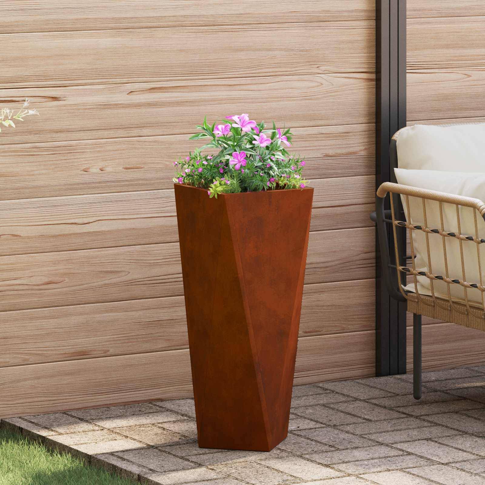 Ein hoher rostfarbener Blumenkübel mit rosa Blumen steht auf einer gekachelten Terrasse neben einem Korbstuhl vor einer hölzernen Wandkulisse.