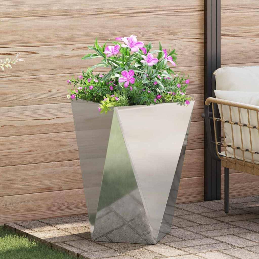 Ein schlanker, eckiger Metallblumentopf beherbergt üppiges grünes Laub mit leuchtend pinkfarbenen Blüten, platziert auf einer Terrasse neben einer Holzwand und Outdoor-Möbeln.