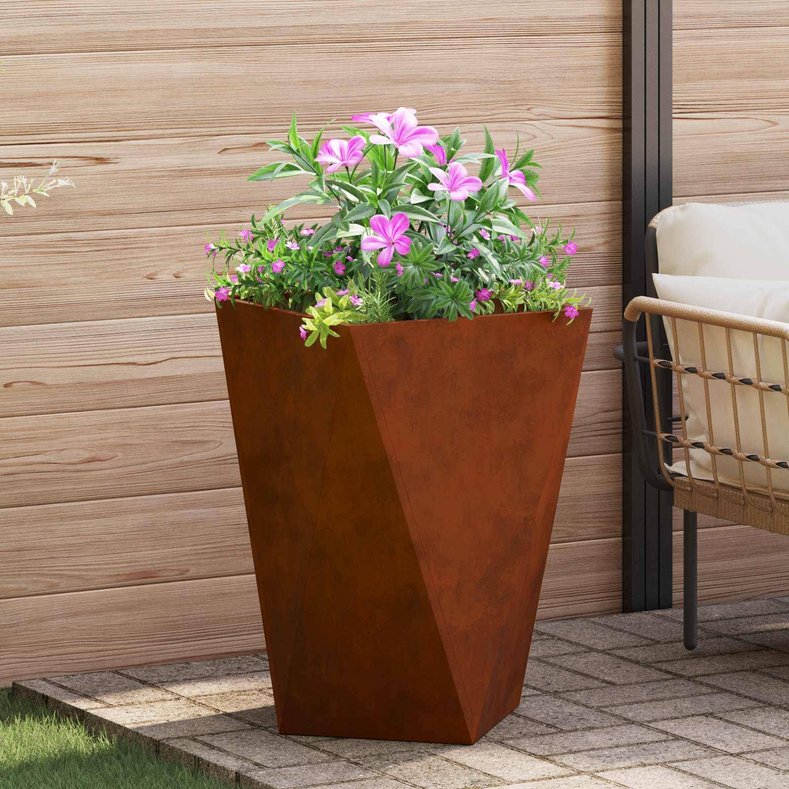 Ein rostfarbener, geometrischer Pflanzkübel mit üppig grünen Pflanzen und rosa Blüten steht auf einer Terrasse mit Holzverkleidung und Gartenmöbeln.