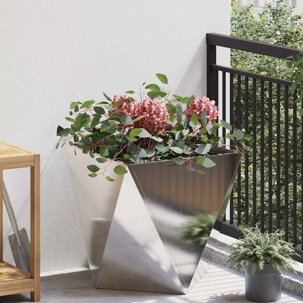 Ein moderner, metallischer dreieckiger Pflanzenbehälter trägt grüne, belaubte Pflanzen und rosa Blumen auf einem Balkon mit einem schwarzen Metallgeländer und einem Holztisch in der Nähe.