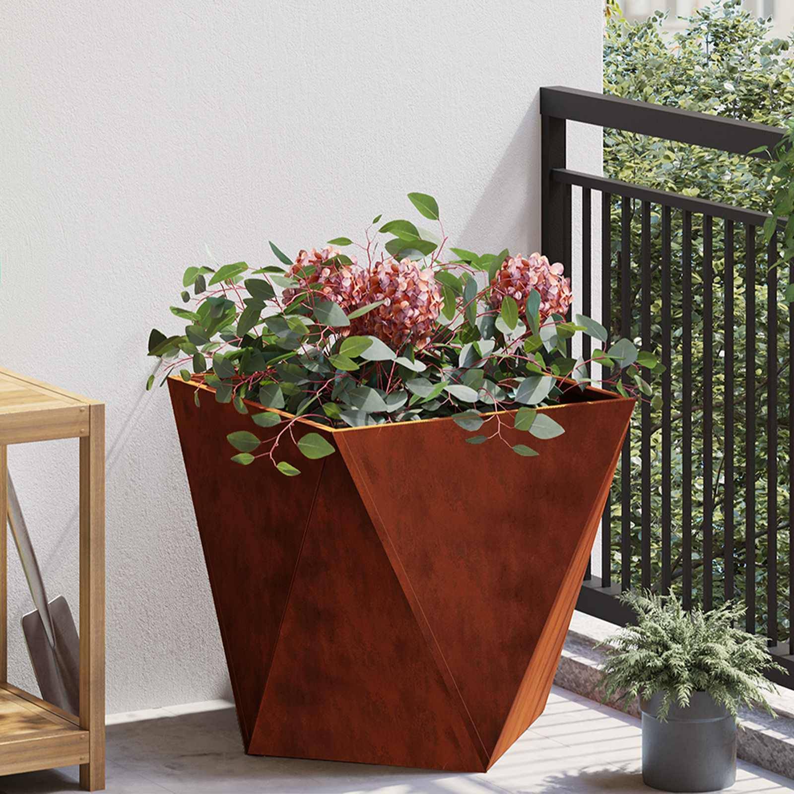 Ein rostfarbener geometrischer Blumentopf mit rosa Blüten und Grünpflanzen steht auf einer Terrasse neben einem schwarzen Geländer und einem hölzernen Regal.