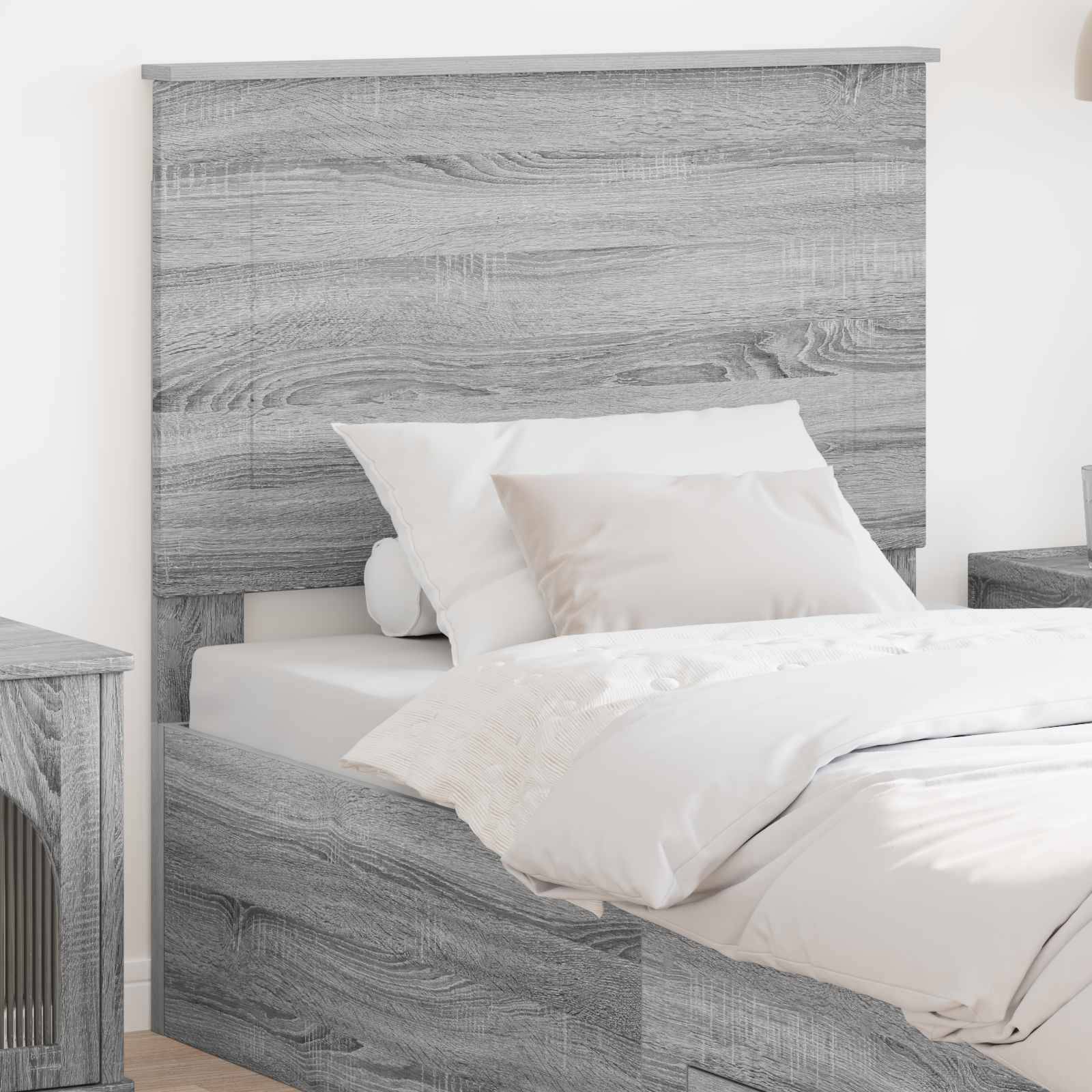 Ein graues Holzbett mit einfachem Kopfteil ist mit weißen Kissen und einer Bettdecke gemacht. Ein Nachttisch ist teilweise auf der linken Seite sichtbar.