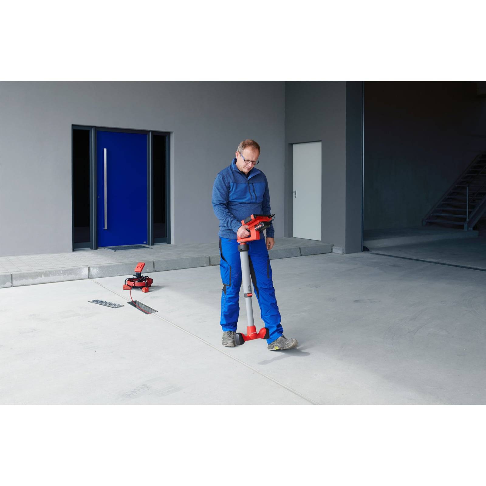 Eine Person in blauer Arbeitskleidung benutzt ein rotes und silbernes Arbeitsgerät in einem modernen Industriegebäude, wobei eine blaue Tür im Hintergrund zu sehen ist.