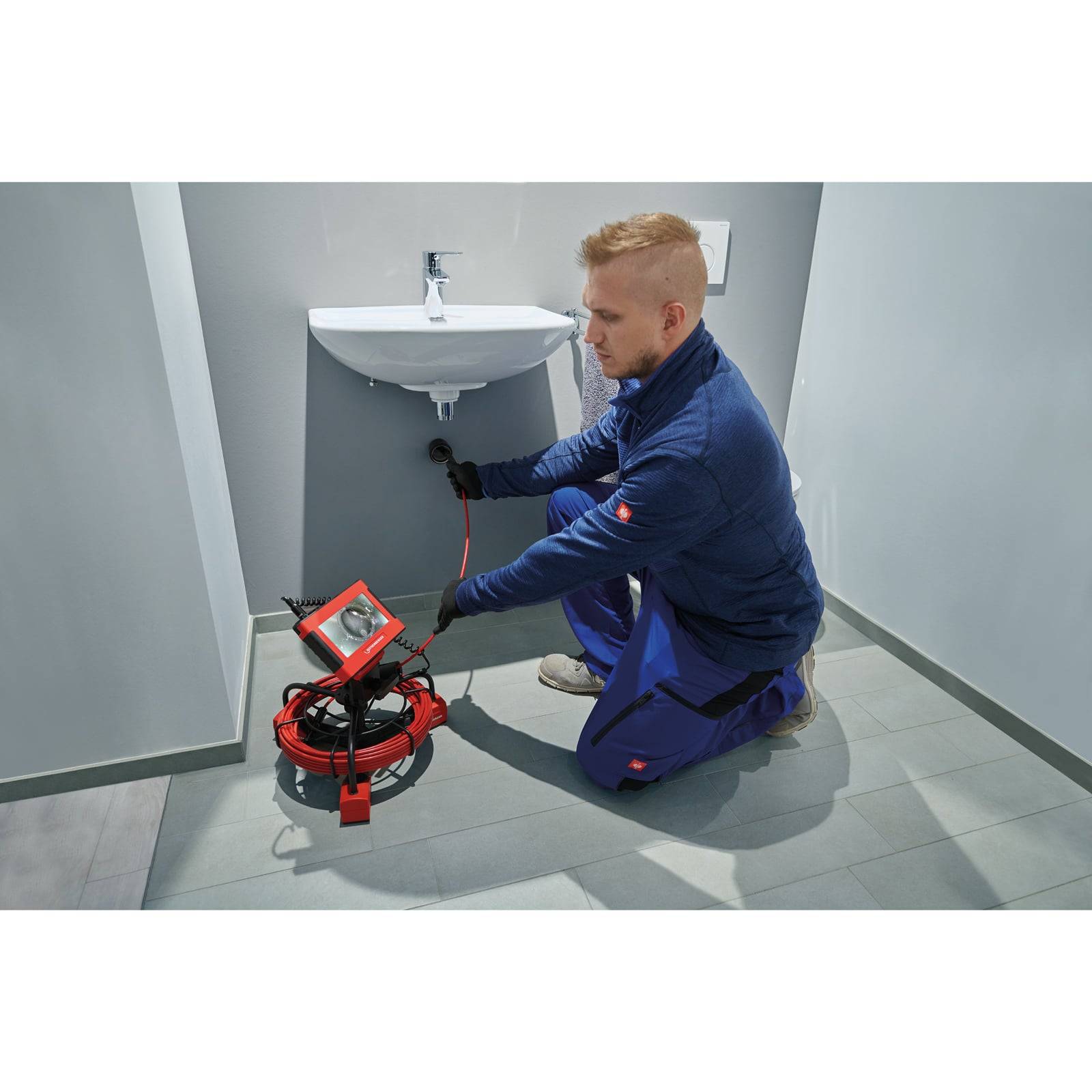 Ein Klempner verwendet ein Gerät zur Inspektion der Rohre unter einem Waschbecken in einem modernen Badezimmer. Er trägt eine blaue Arbeitsuniform und Handschuhe und konzentriert sich auf seine Aufgabe.
