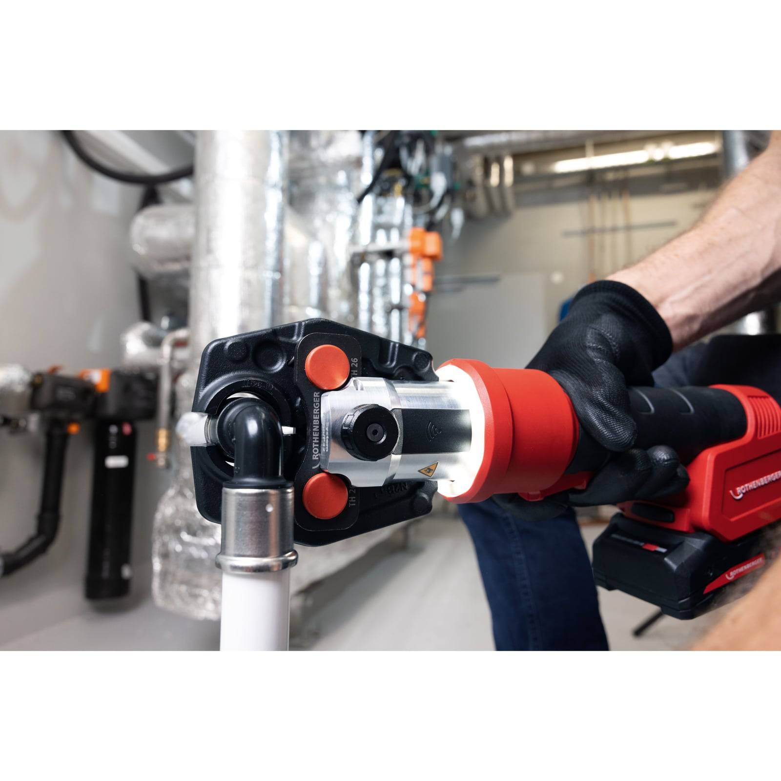 Eine Person verwendet ein rot-schwarzes Arbeitsgerät, um Rohre in einer industriellen Umgebung zu verbinden, umgeben von metallischer Infrastruktur.