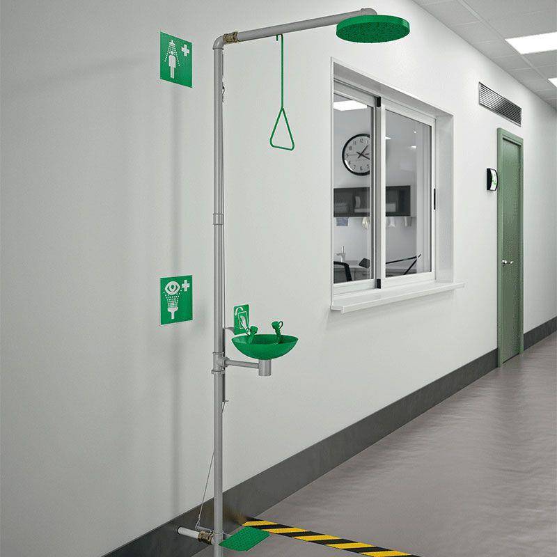 Notfall-Augenspülstation mit grünem Duschkopf und Handwaschbecken an der Hallenwand außerhalb eines Klinik- oder Laborraums montiert.