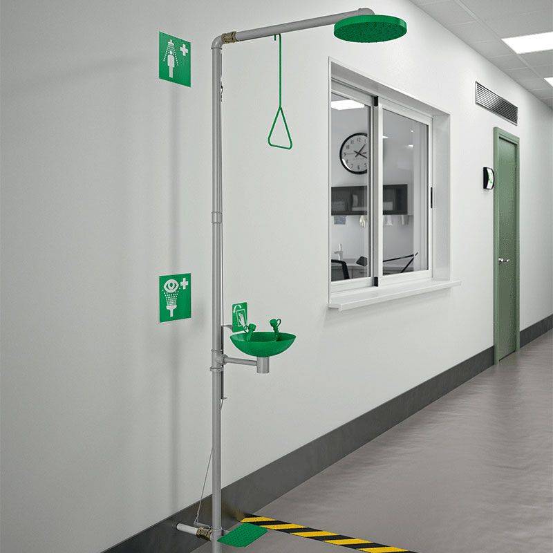 Notdusche und Augenspülstation in einem Flur nahe einem Fenster, gekennzeichnet mit grünen und weißen Sicherheitsschildern für einen schnellen Zugang.