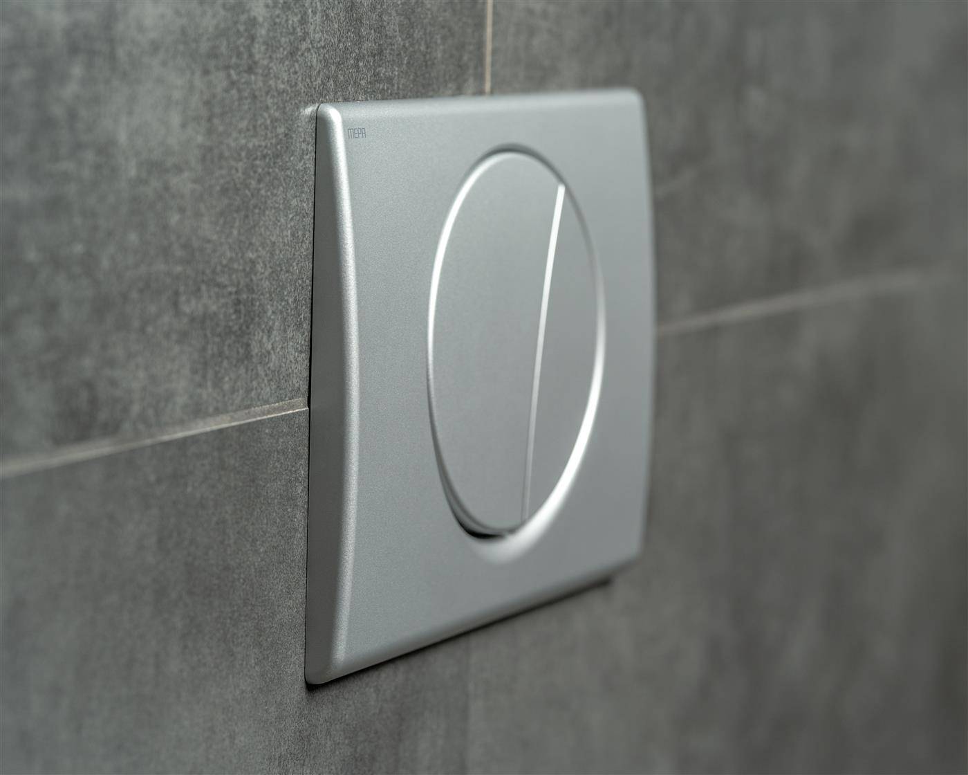 Silberner Dual-Flush-Toilettenknopf an der Wand auf einer dunkelgrauen gefliesten Badezimmerwand.