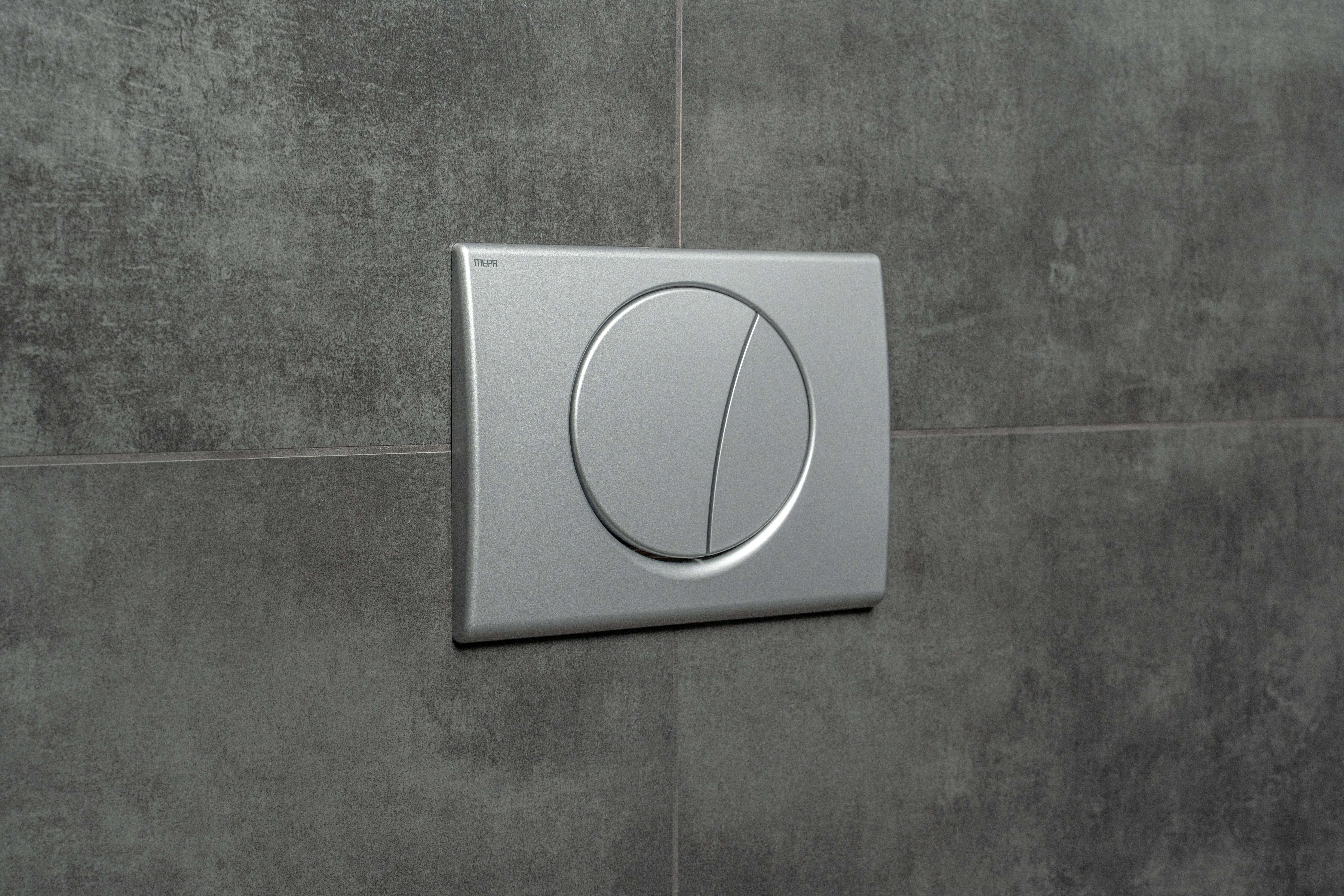 Ein silberner Dual-Flush-Toilettenknopf, der an einer gefliesten Wand montiert ist und wassersparende Spüloptionen mit einem eleganten, modernen Design bietet.