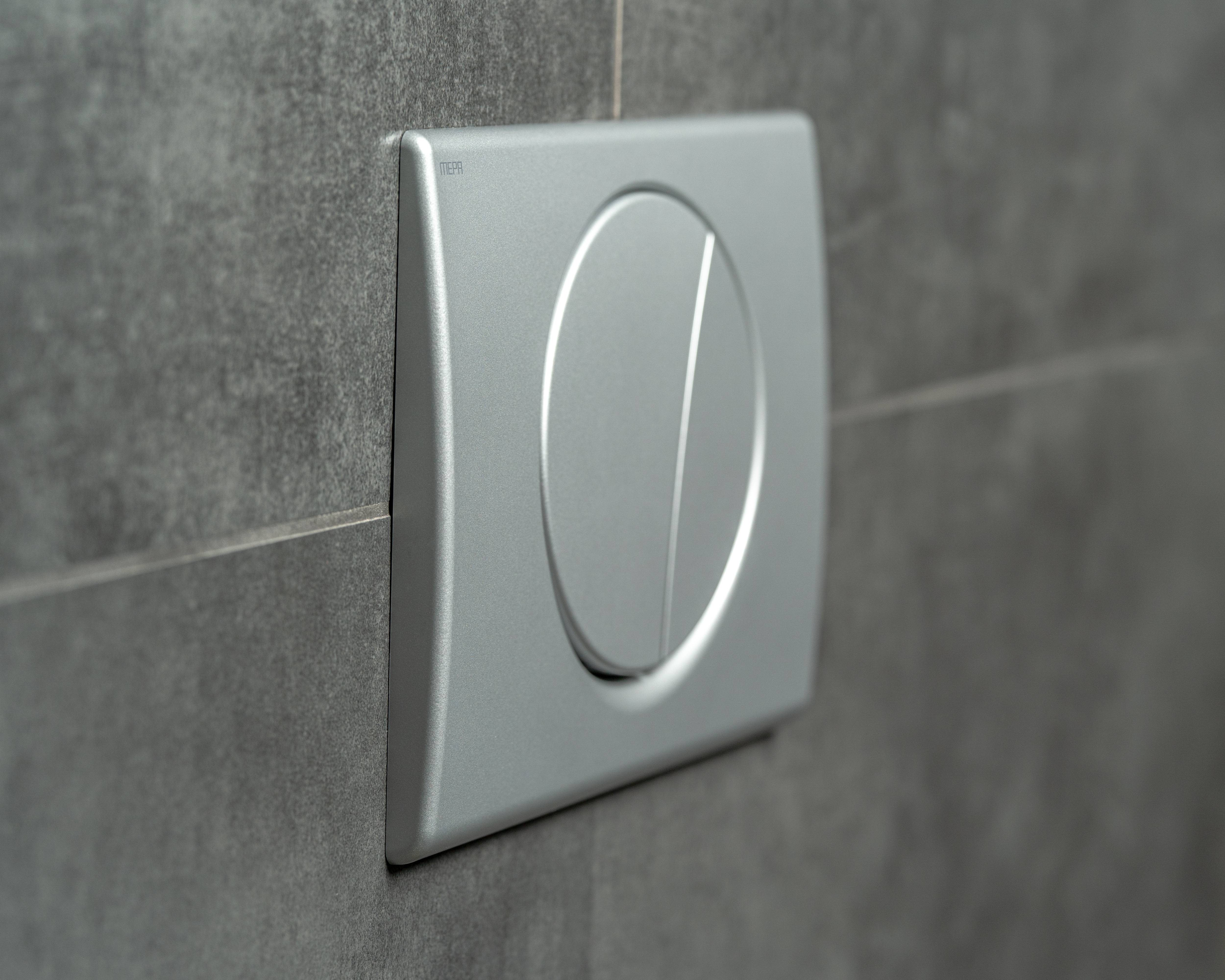 Ein silberner Dual-Flush-Toilettenspülknopf an einer grauen gefliesten Wand mit modernem, ovalen Design.