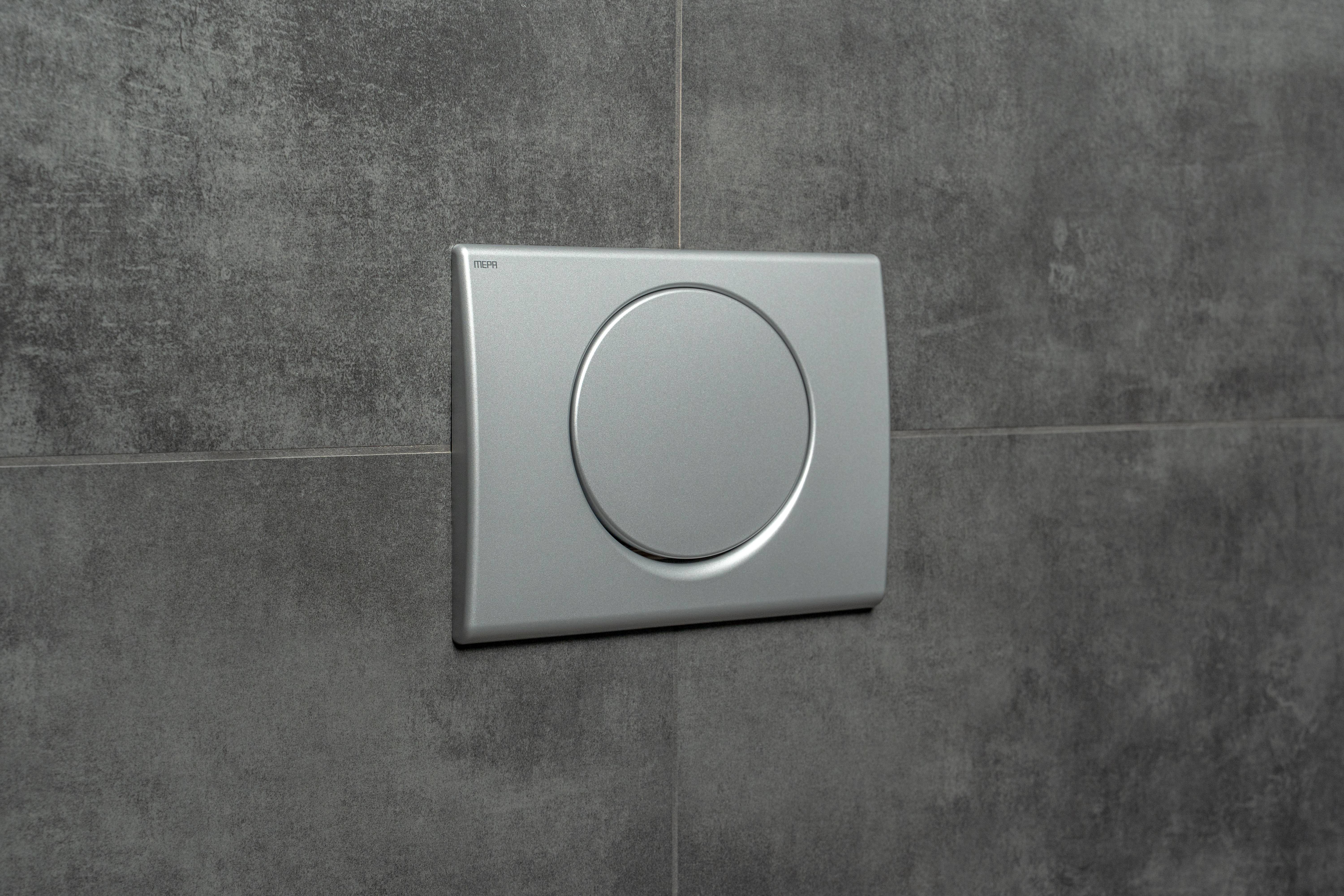 Eine Edelstahl-Betätigungsplatte mit modernem kreisförmigem Design ist an einer grauen gefliesten Badezimmerwand montiert.