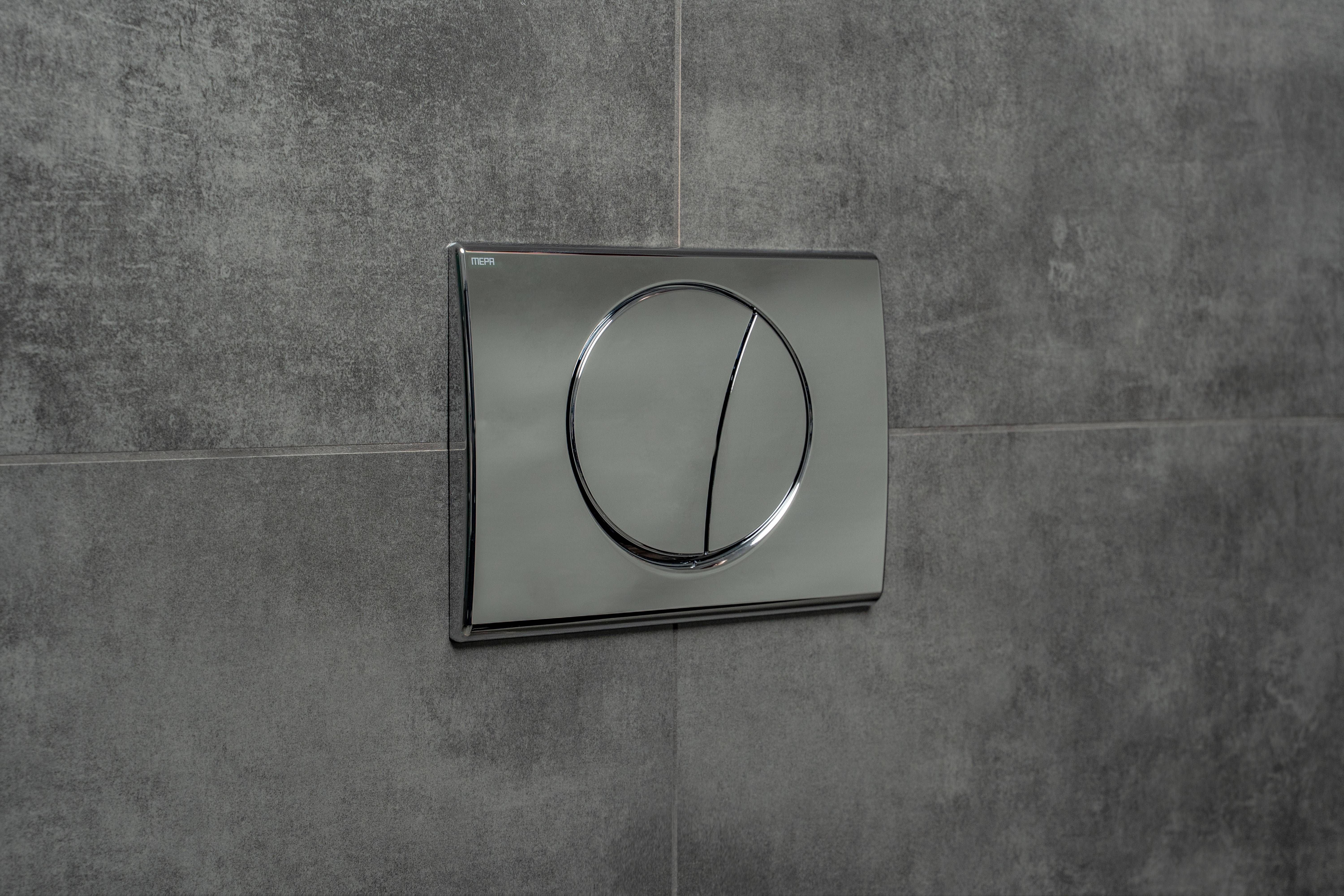 Eine glänzende Metallfläche in Form einer Einbauplatte an einer grauen gefliesten Wand mit zwei Tasten für unterschiedliche Spüloptionen.